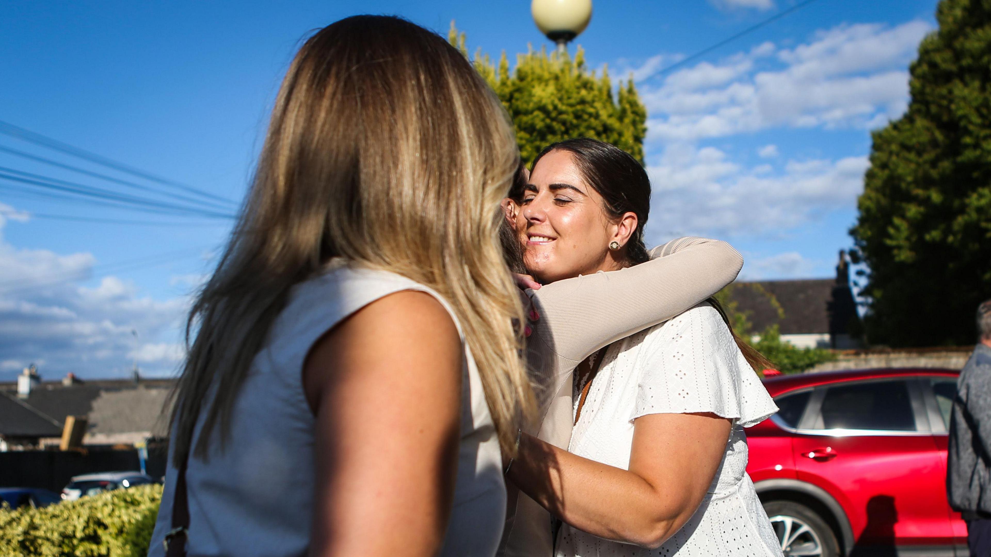 A woman with long blonde hair stands in front of two women hugging each other. 