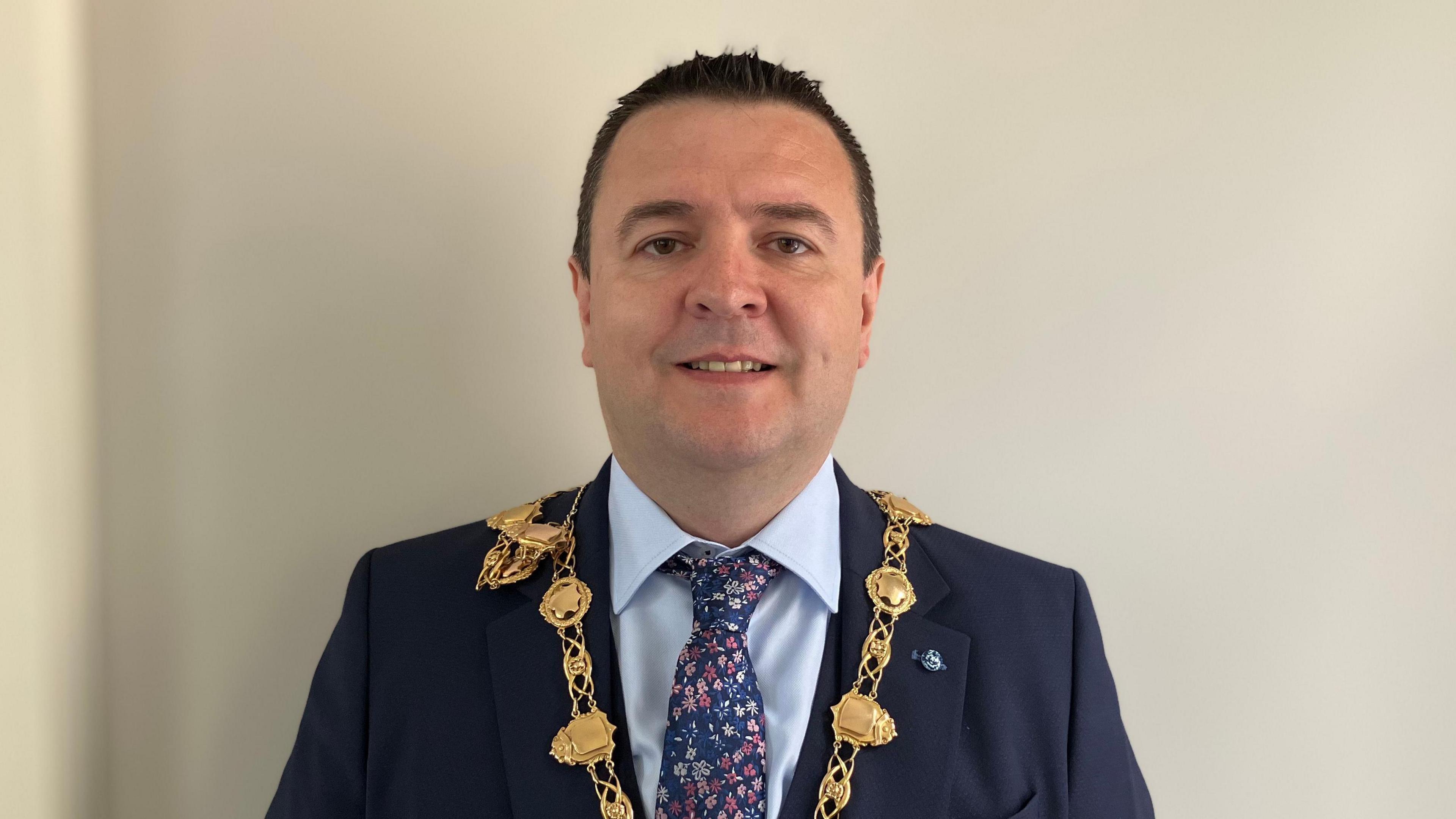 Derry mayor Ruairí Mc Hugh looking directly at the camera and smiling.  He has very short, dark hair and is wearing a navy suit, pale blue shirt, navy patterned tie and his gold chain of office. 