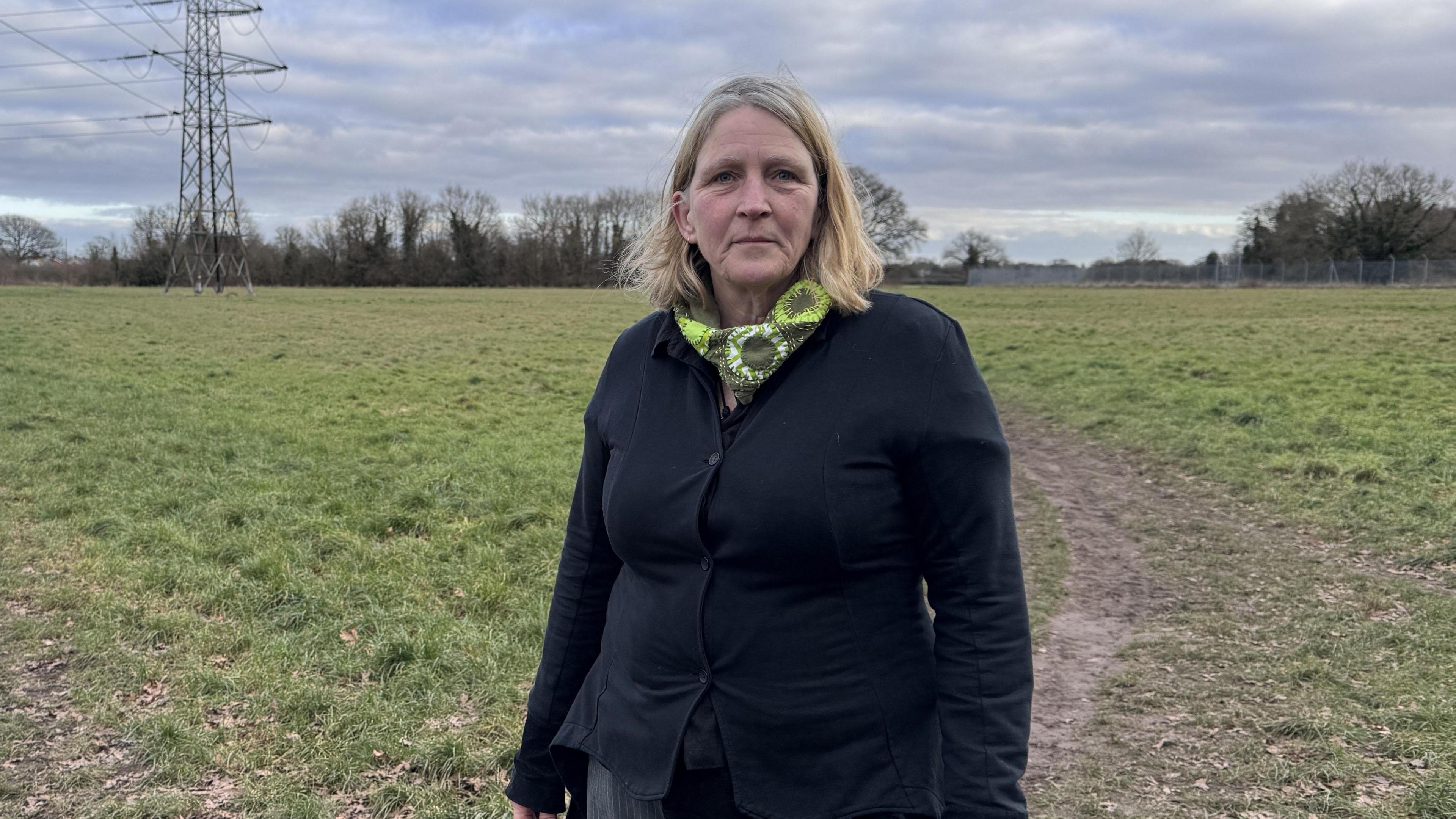 Andrea Luxford Vaughan is a blonde woman wearing a navy cardigan and a green scarf. She is standing on the Middlewick site and looking at the camera.