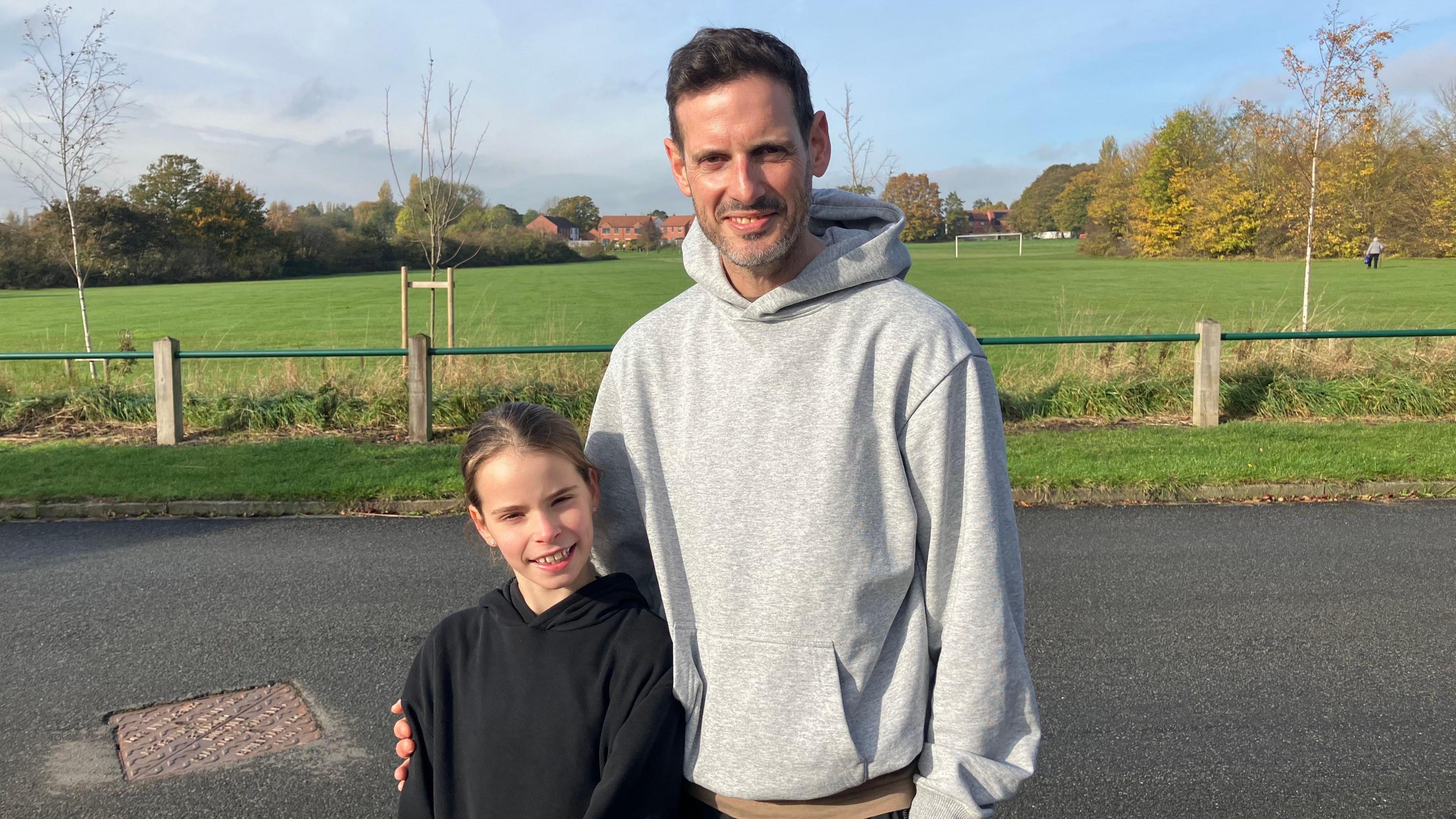 A young girl with brown hair stands next to her father, who also has dark hair, with a grass field in the background.