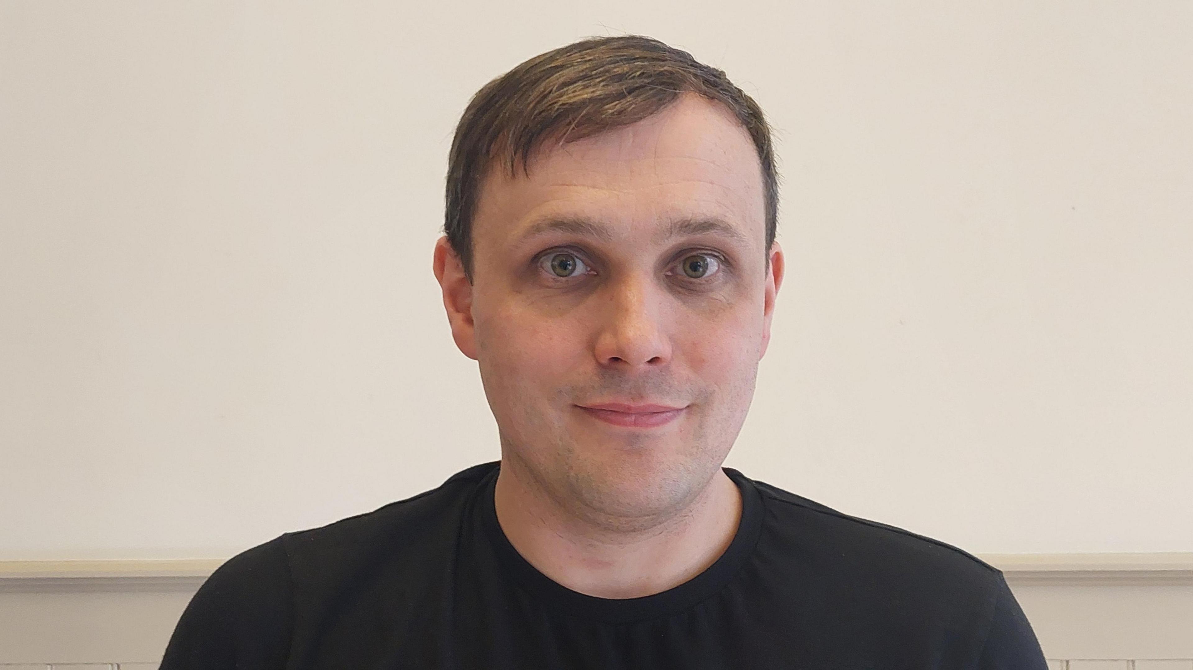 Darren Murphy has short brown hair and is wearing a black T-shirt. He is standing against a light-colored wall with wooden paneling.