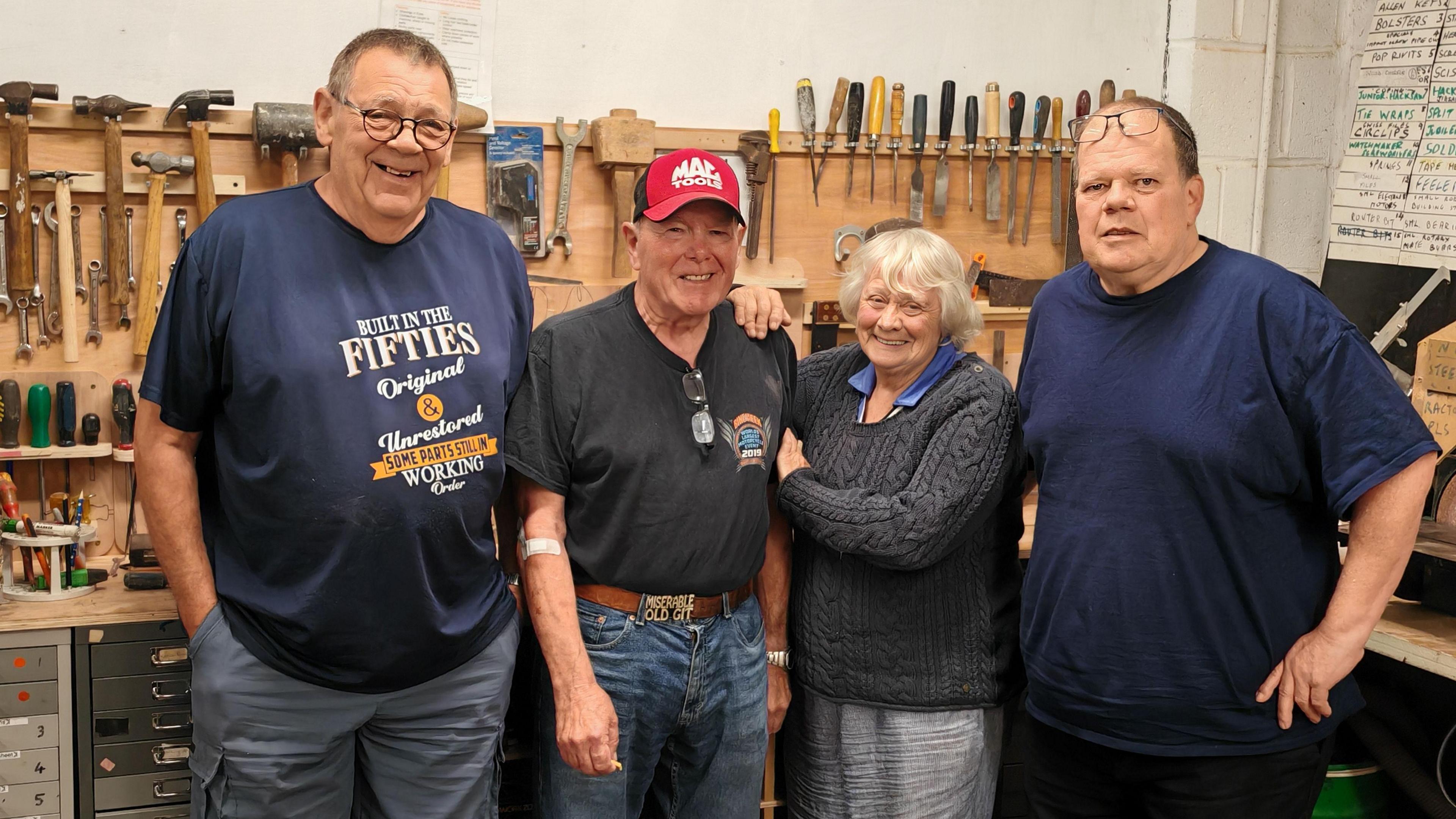 Rick Callingham, Syd McDonald, Ann Pritchard, Steve Barton stand shoulder to shoulder in the Spelthorne Shed, with racks of tools in the background. The men all wear t-shirts and jeans, while Ann wears a blue blouse and grey cable knit sweater.