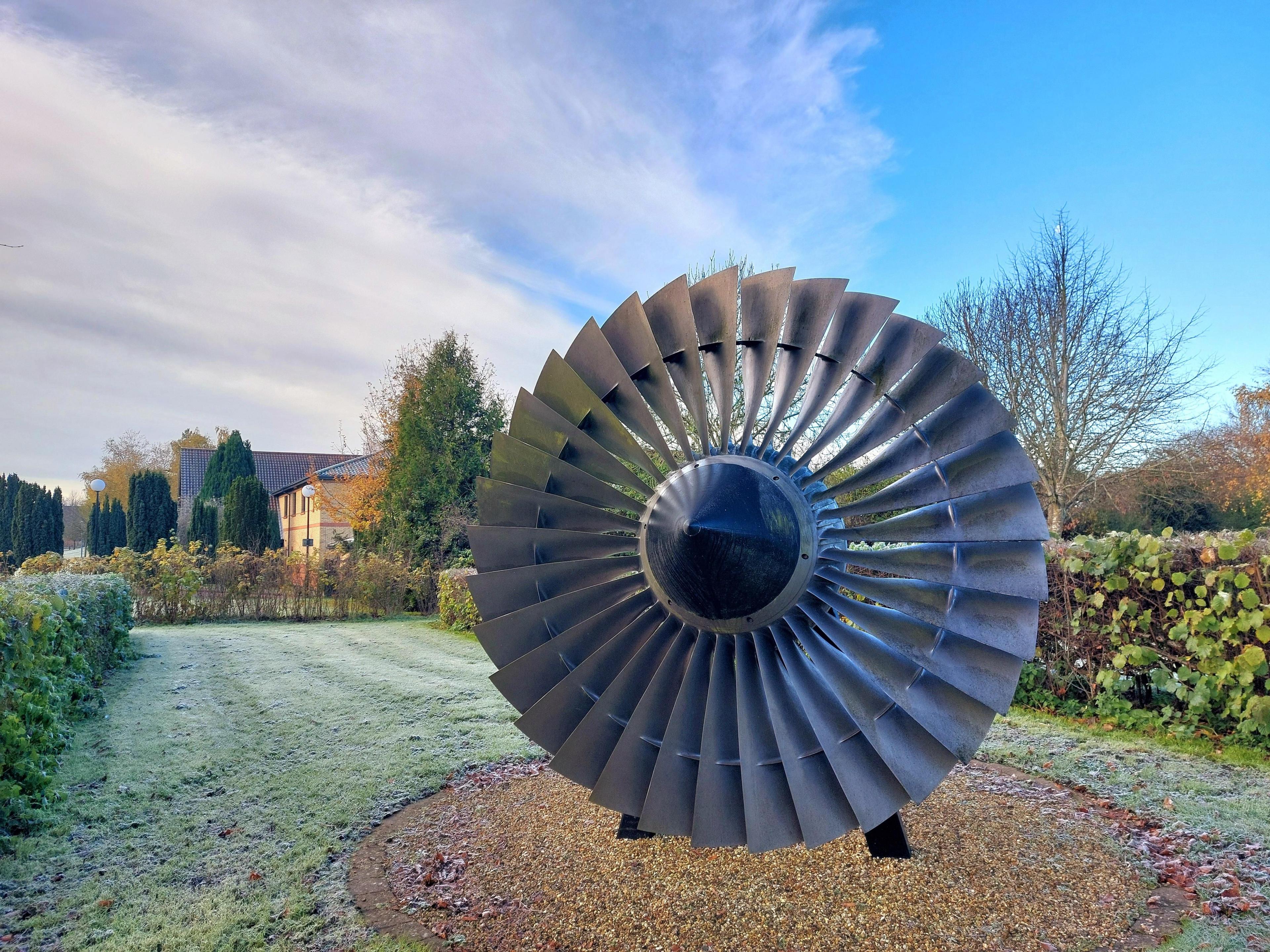 An aircraft fan is placed on a wooden stand on a patch of gravel in the middle of a lawn with box hedges either side and campus buildings in the background. There is frost on the grass. 