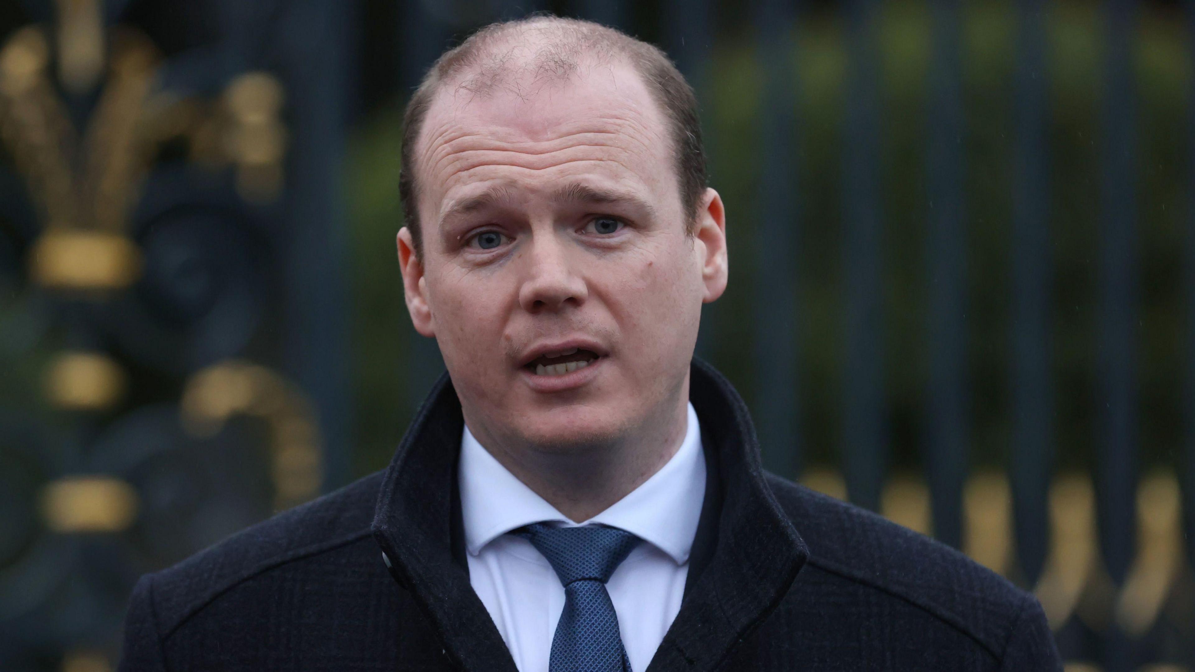 Minister for Communities, Gordon Lyons wearing a dark woollen coat, white shirt and navy tie.