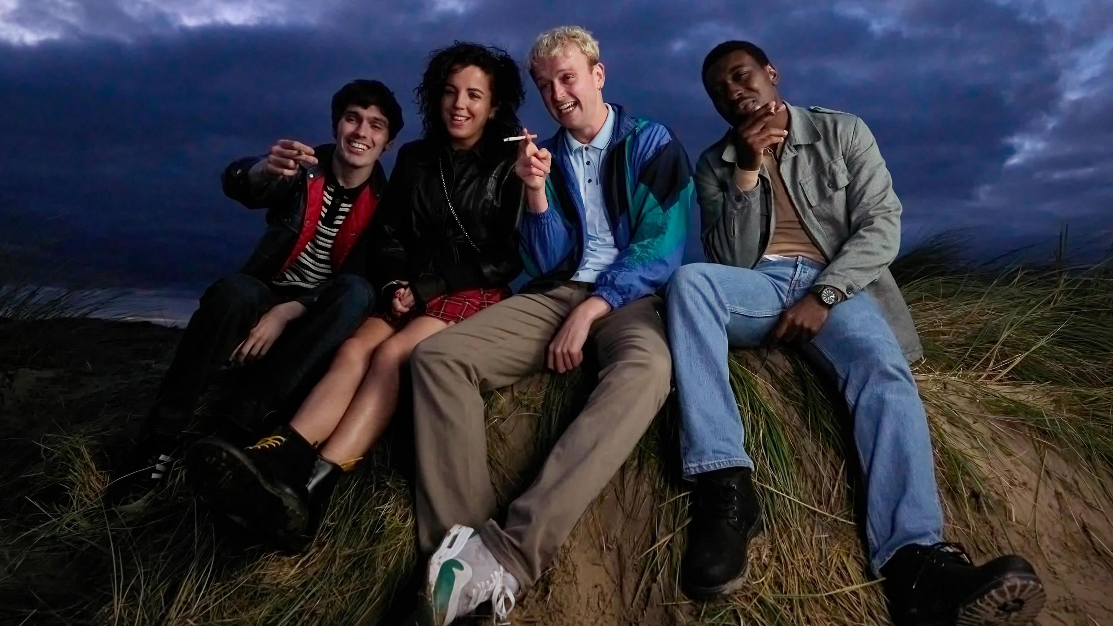 Four friends sit smiling and smoking on a grassy mound in a rural field. Clouds roll through a dark sky overhead. 