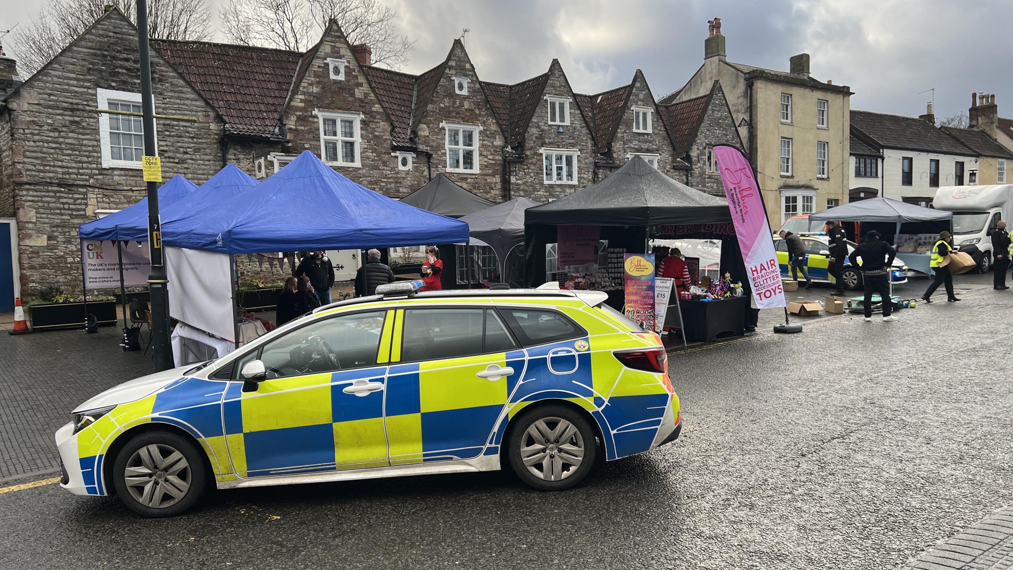 A police car parked in the middle of the road. Behind it are several stalls set up for the market, in front of a historic looking stone building. 