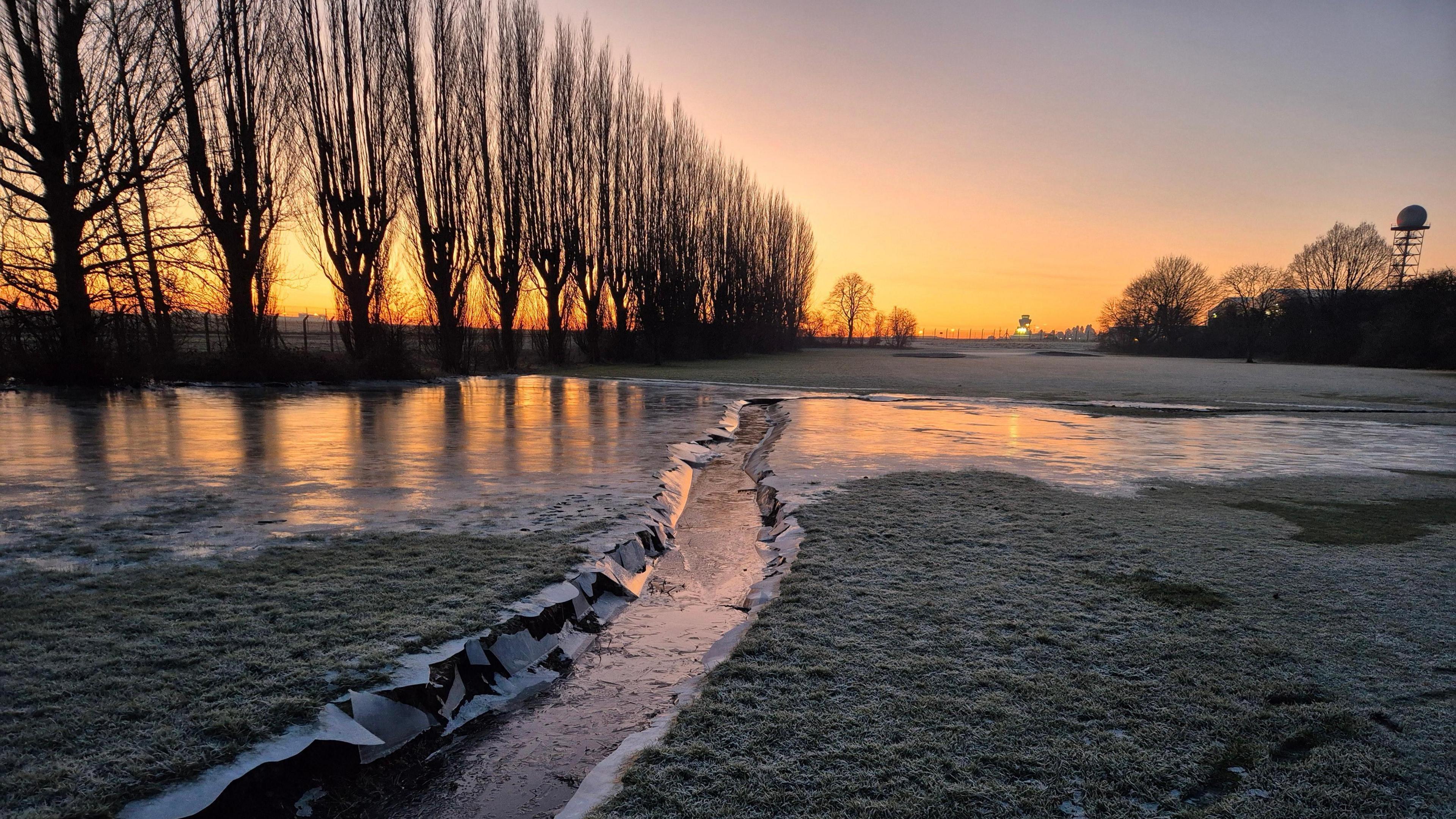 A stream is frozen over and surrounded by green grass that is also frozen. A line of trees sit to the left of the field. An orange and red sunrise is seen in the sky and reflects off the frozen water.