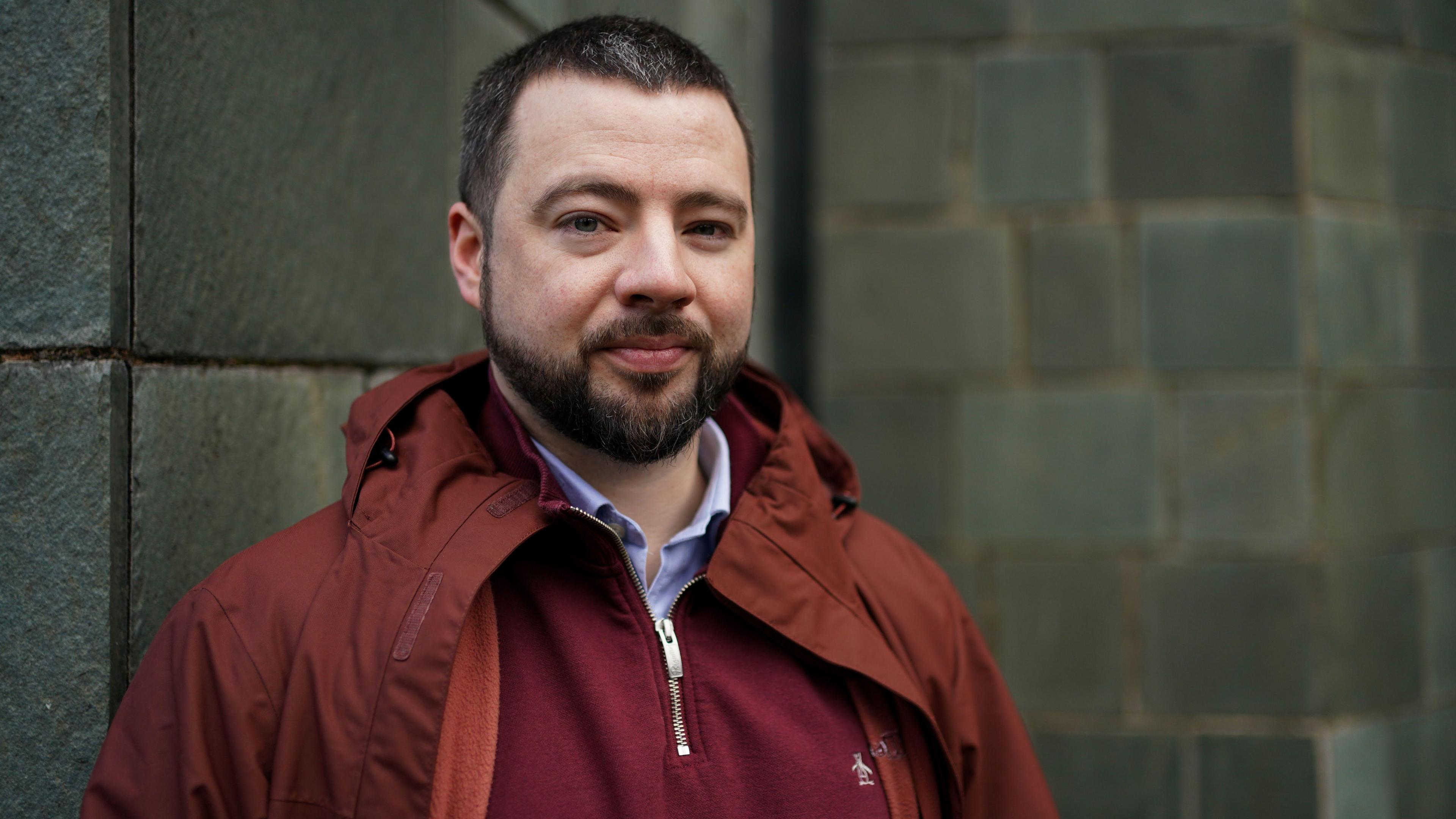 Jason Evans in a red coat, three-quarter jumper and a shirt. He has a beard and is looking into the camera. Behind him is a grey brick wall.