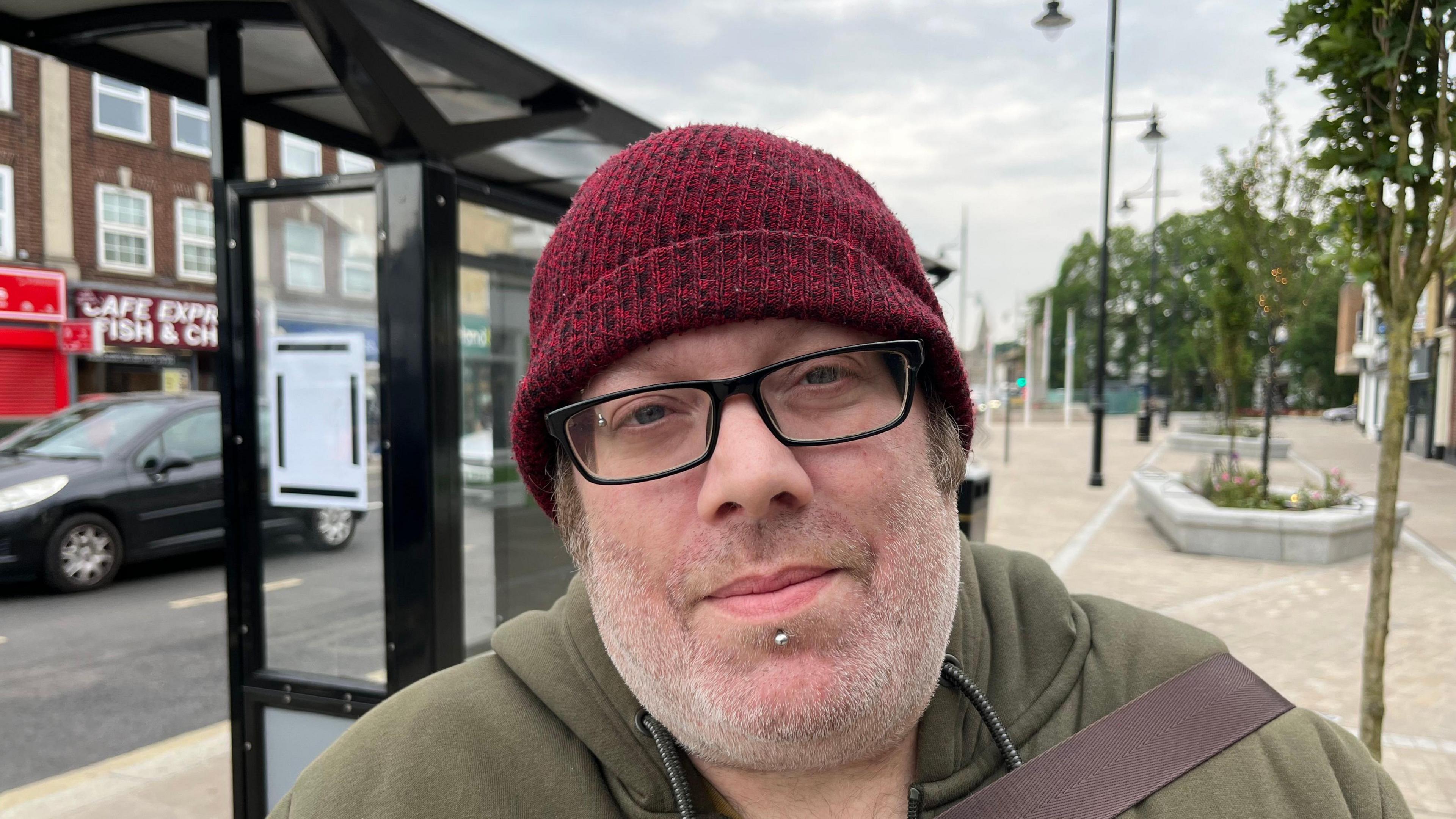 Craig Denyer, 43, has stubble on his chin and a dark red bobble hat on his head. He has glasses with dark frames and a gold stud just under his bottom lip. He is wearing a khaki hoodie with the strap of a bag over his shoulder. Behind him is a bus stop with cars passing on the road. 