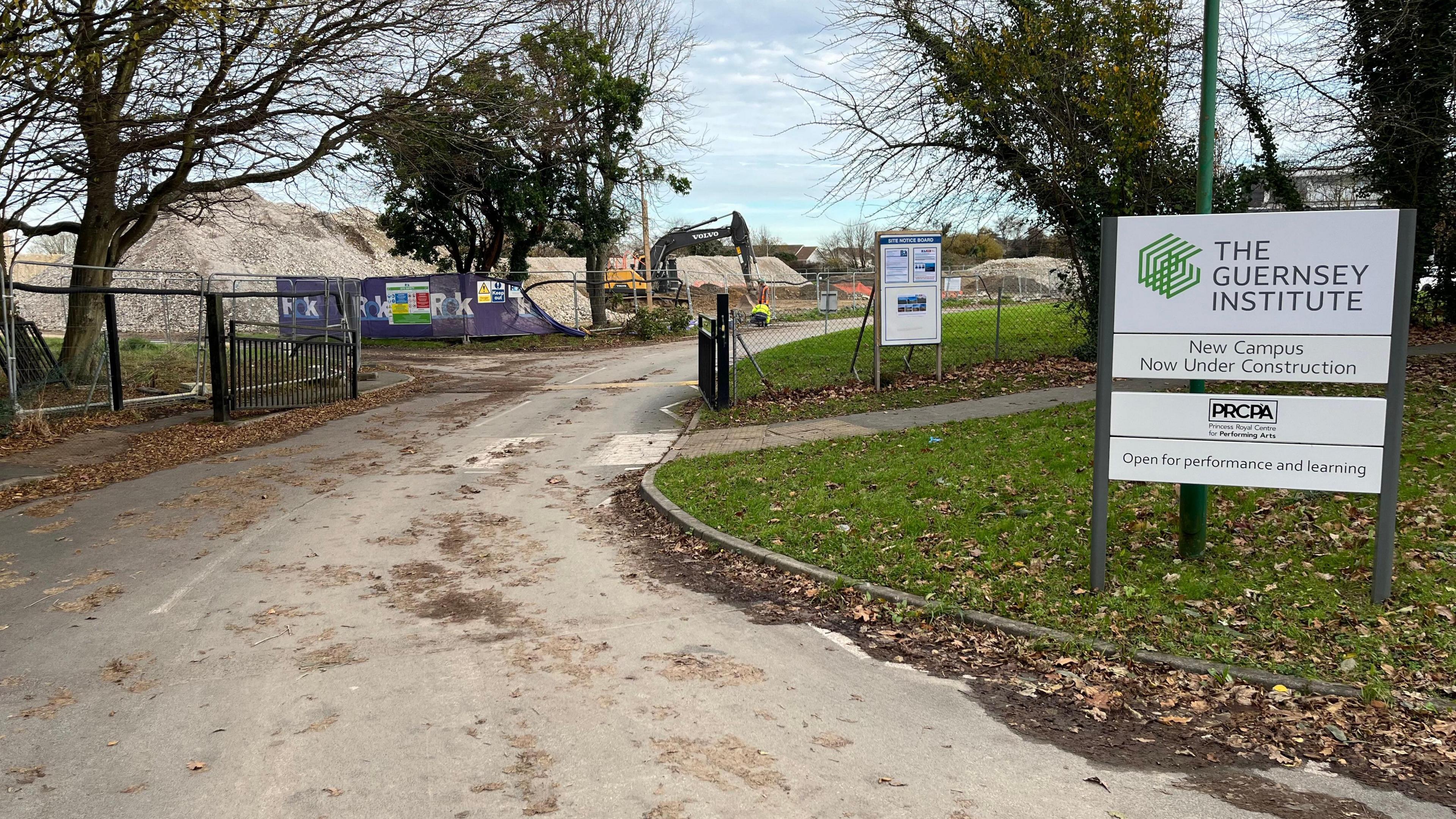 A white sign which says The Guernsey Institute, with new campus under construction underneath. To the left of the sign is a gate and behind it are large mounds of rubble and a large digger. 