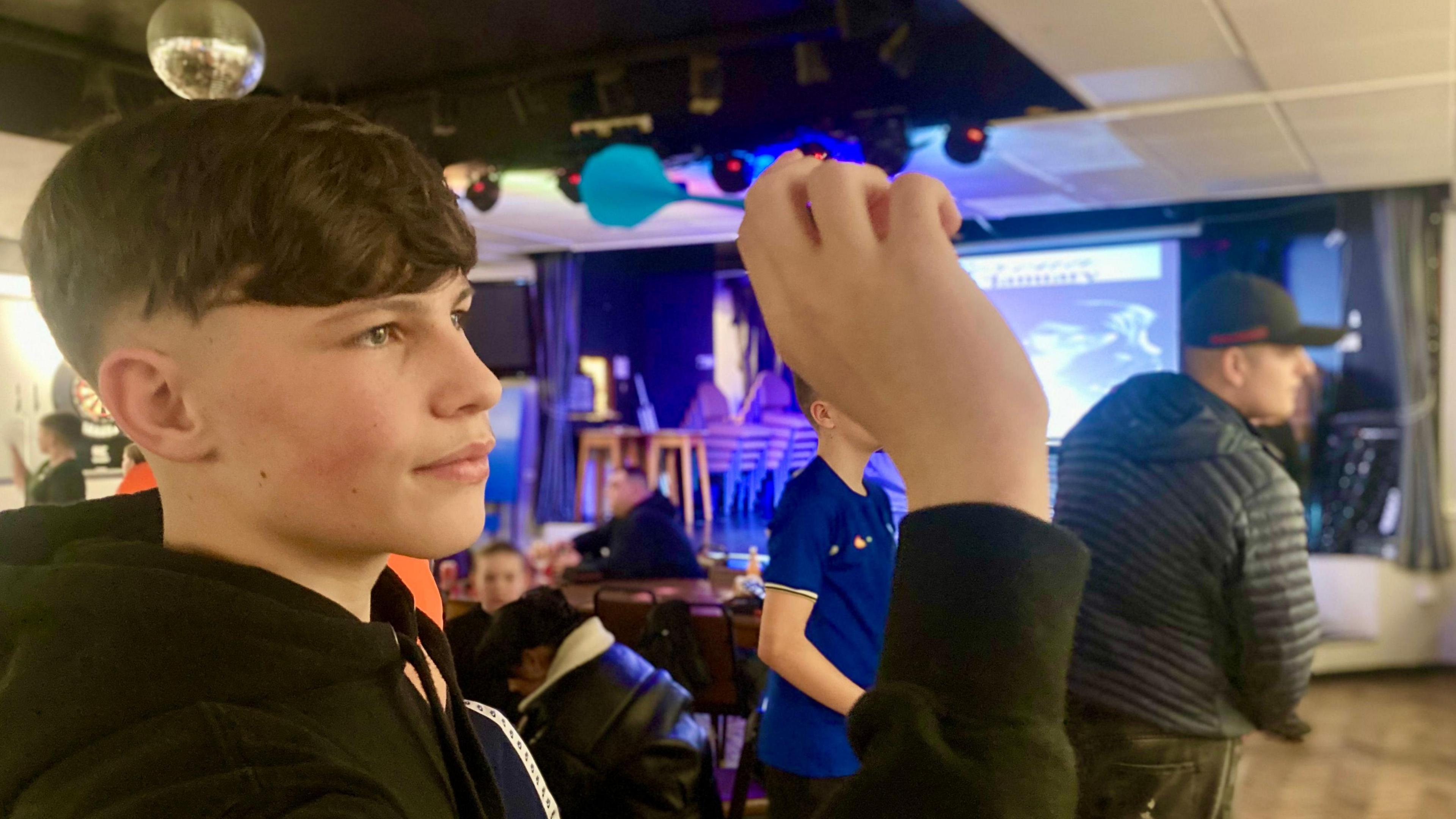 Rylie, a 15-year-old boy aims a dart at the board. He has short dark hair and is wearing a black hooded top. He is holding the dart in his right hand. It has light blue flights. Behind him are other youngsters playing darts in a club in Hull.