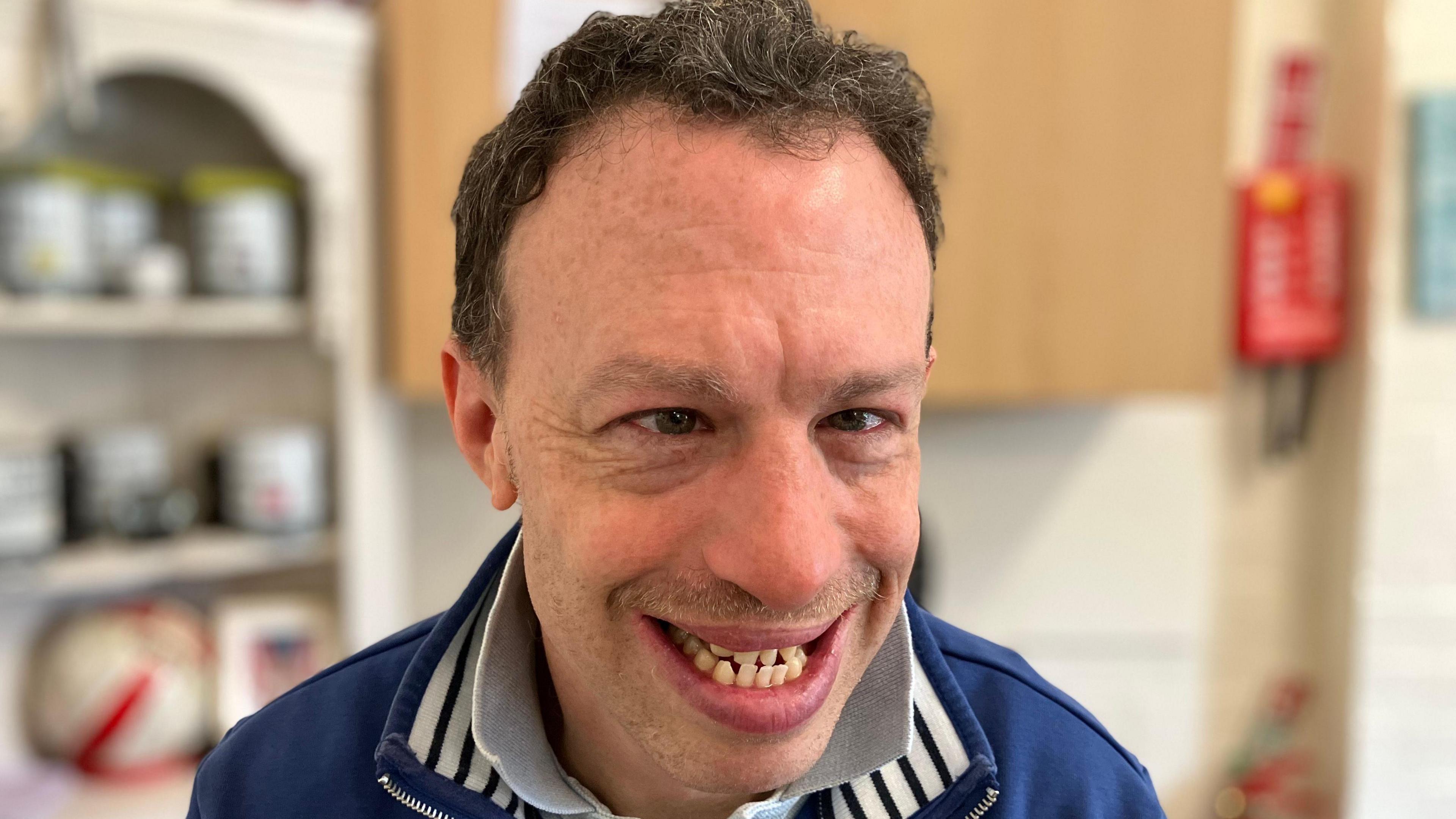 A head and shoulders shot of Ben Greener who is smiling and looking at the camera. He is wearing a collared t-shirt and a blue jumper. He is standing in a kitchen with the background out of focus.