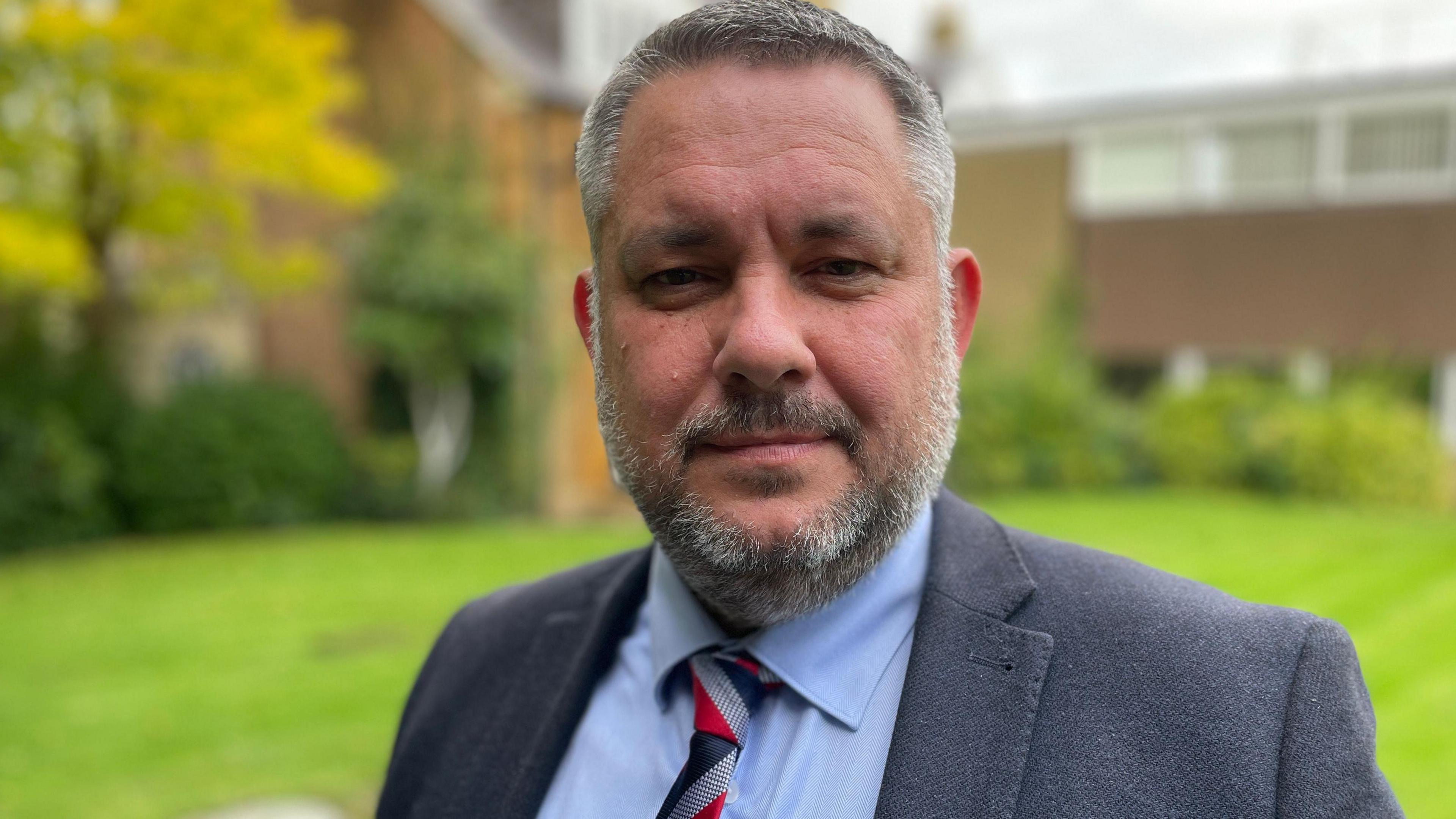 A picture of Conservative Leader of North Northamptonshire Council Jason Smithers. He has short greying hair with a sculpted beard and moustache and is wearing a grey suit jacket and blue shirt with a blue, grey and red tie.