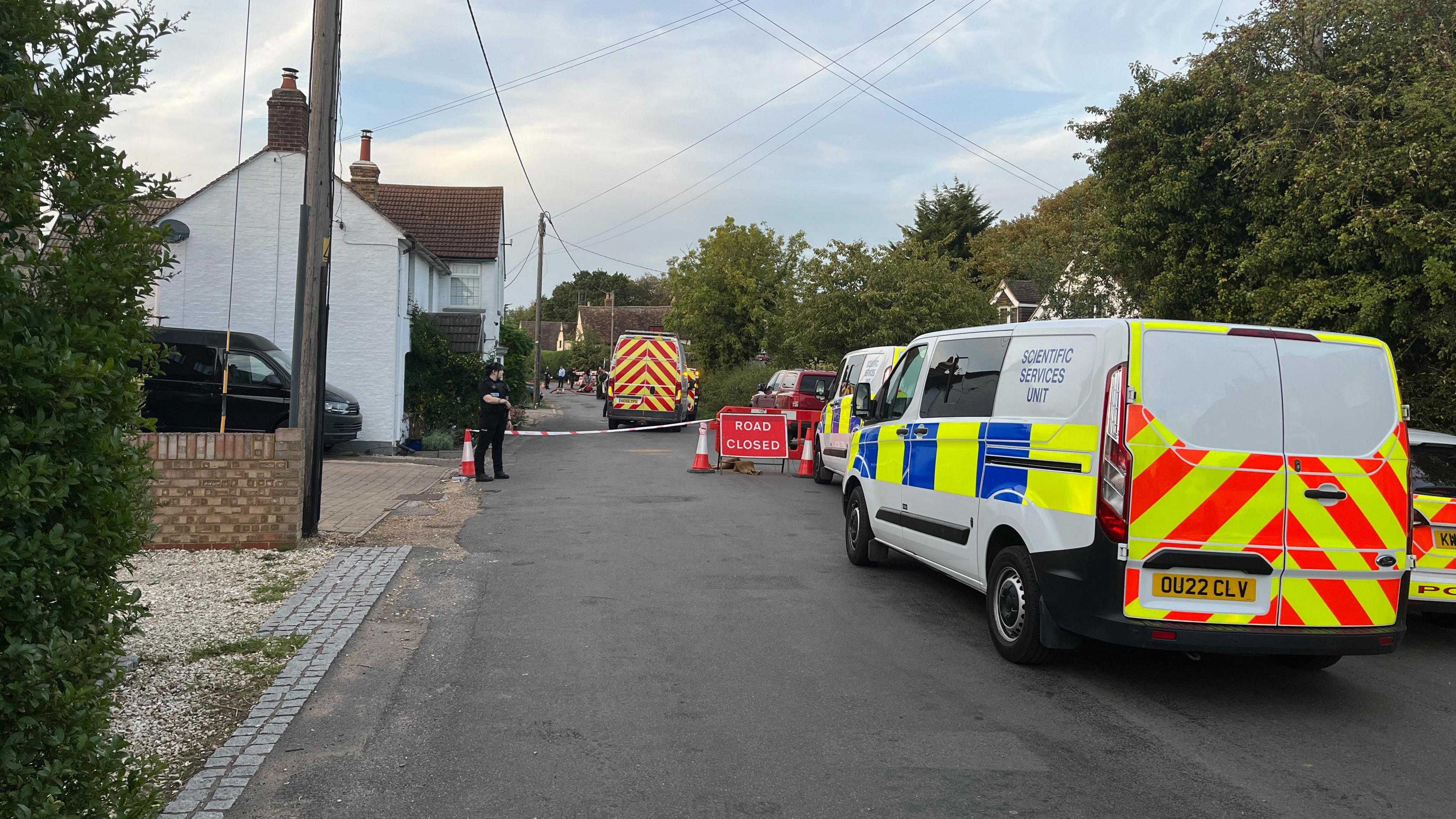 Police vans from the scientific services unit parked on a road, which has houses on the left and trees on the right. There is a road closed sign with a police officer standing next to it.