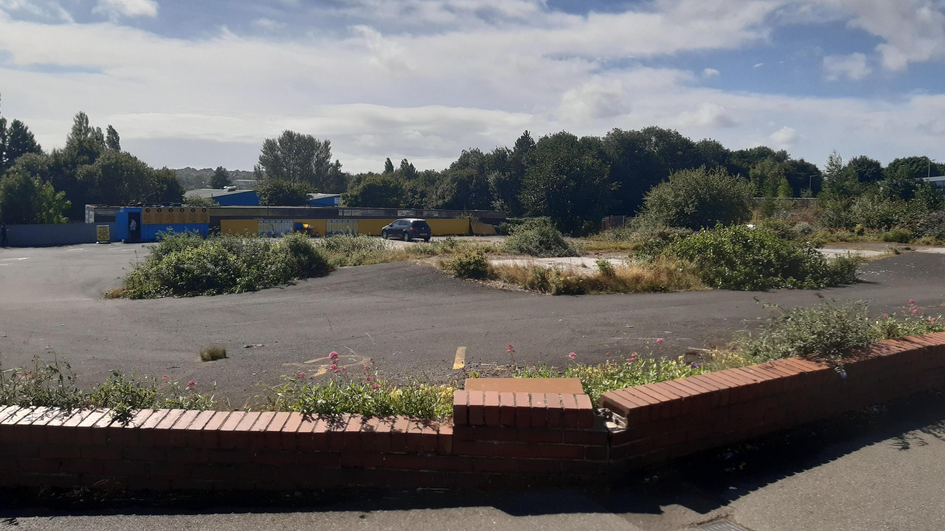 A image of an empty urban site with a low brick wall in the foreground then a concrete forecourt with bushes growing everywhere. 