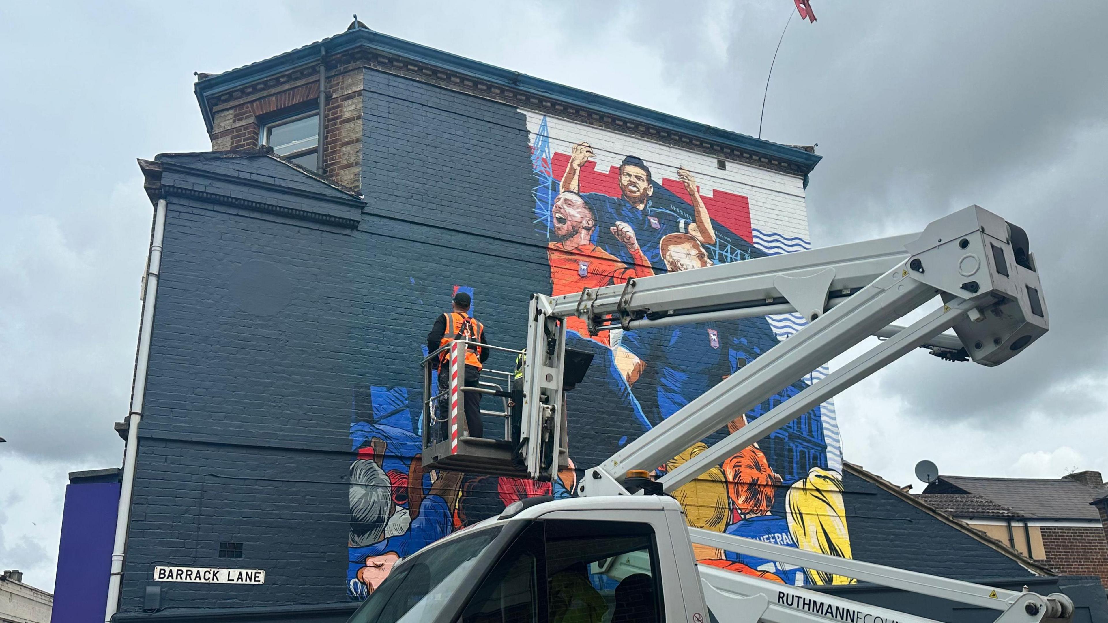 Another angle of the painter on a platform lift paints over a mural that depicted Ipswich Town's players and manager on the side of a building.