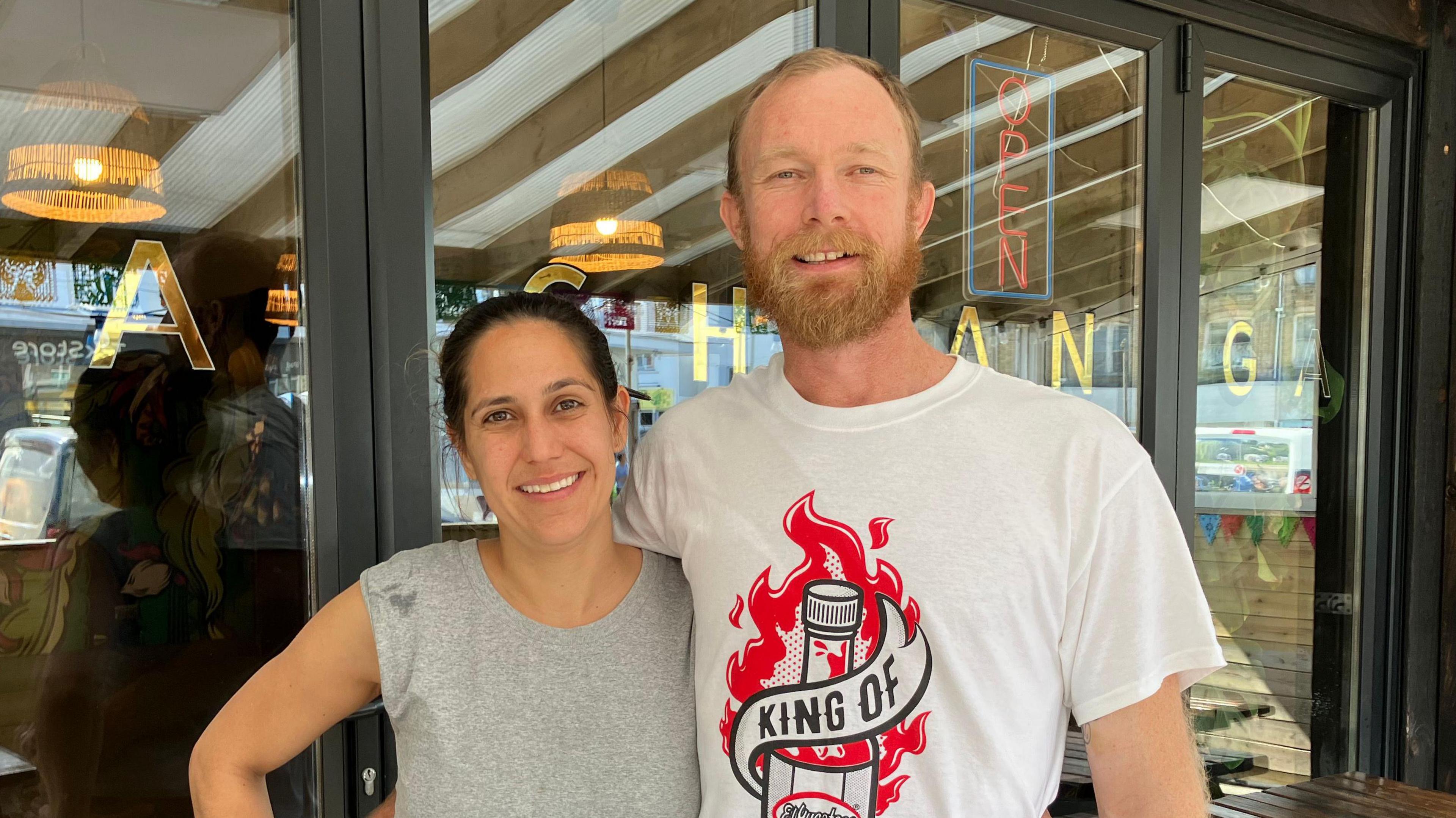 John Dean and wife Carla - both smiling, Carla with black hair tied back wearing a short sleeve grey top, John wearing a white top with a red fire graphic behind a bottle with a wrap saying KING OF, background of glass doors