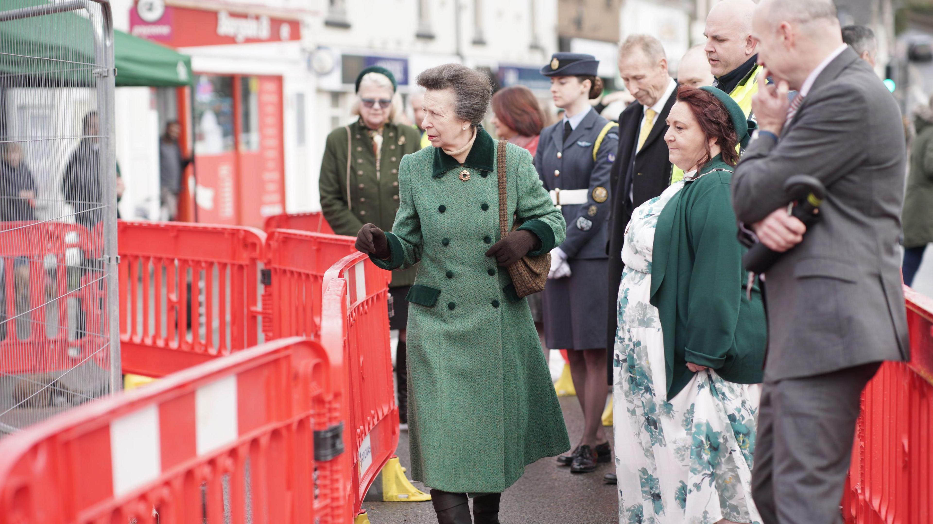 Princess Anne, wearing a long green jacket, brown gloves and black boots, walks down Lydney high street in between a row of red barriers. She is joined by a number of other people. 