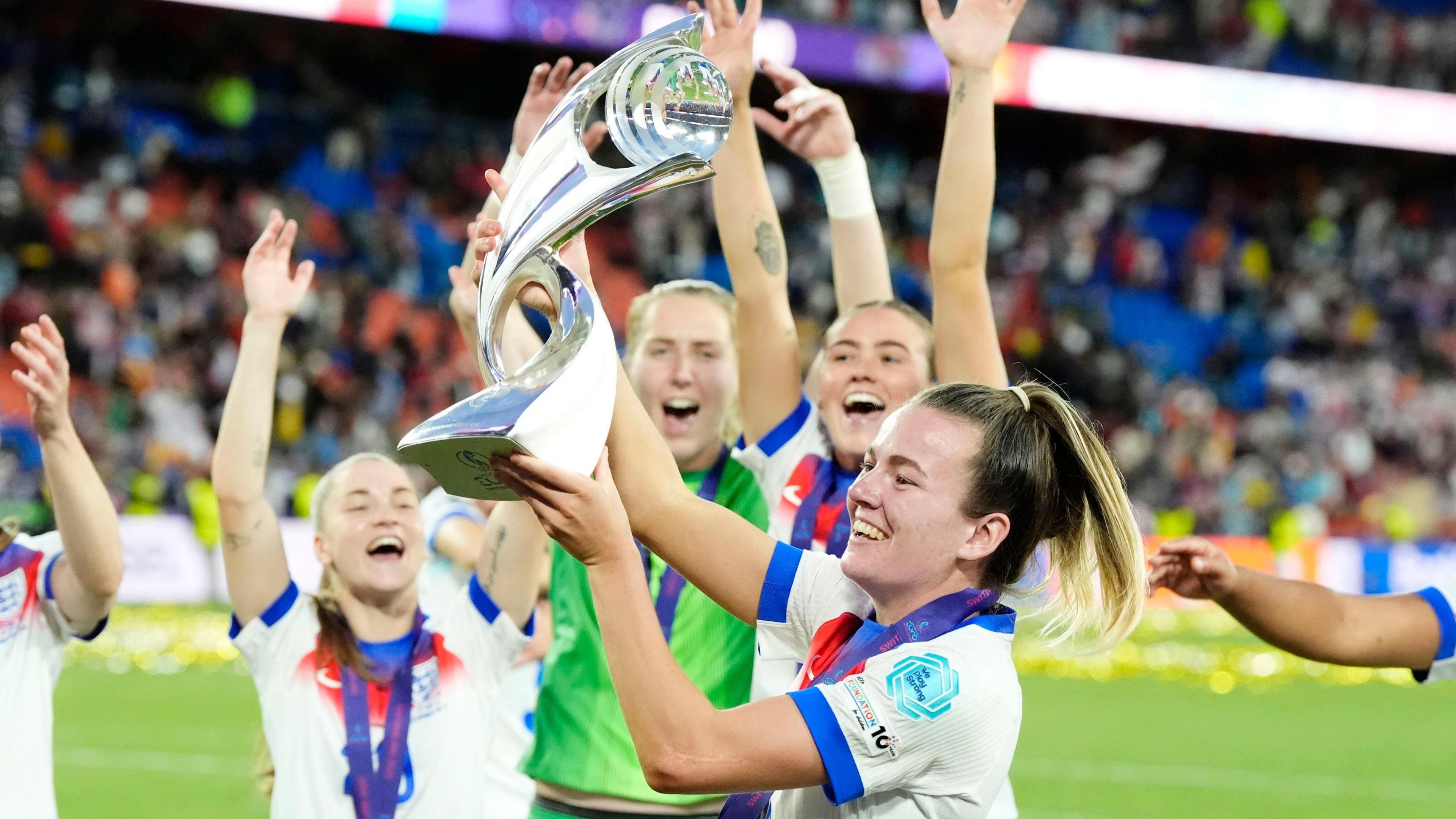 A female football player lifts a trophy in celebration as she smiles with her team-mates celebrating around her