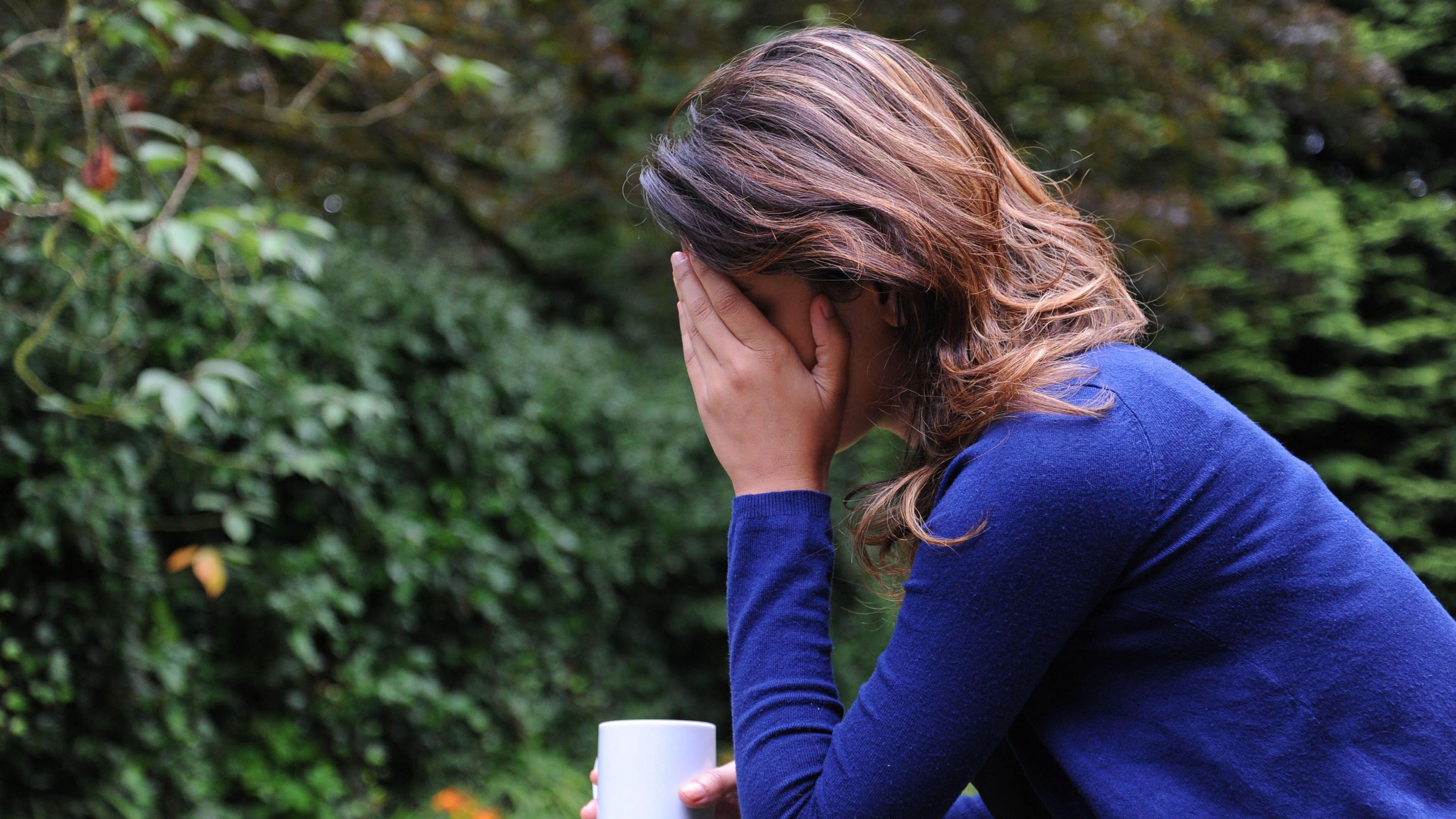 A  picture of a woman holding one hand to her head. In the other hand she is holding a cup. There is greenery behind her as if she is sitting in a garden.