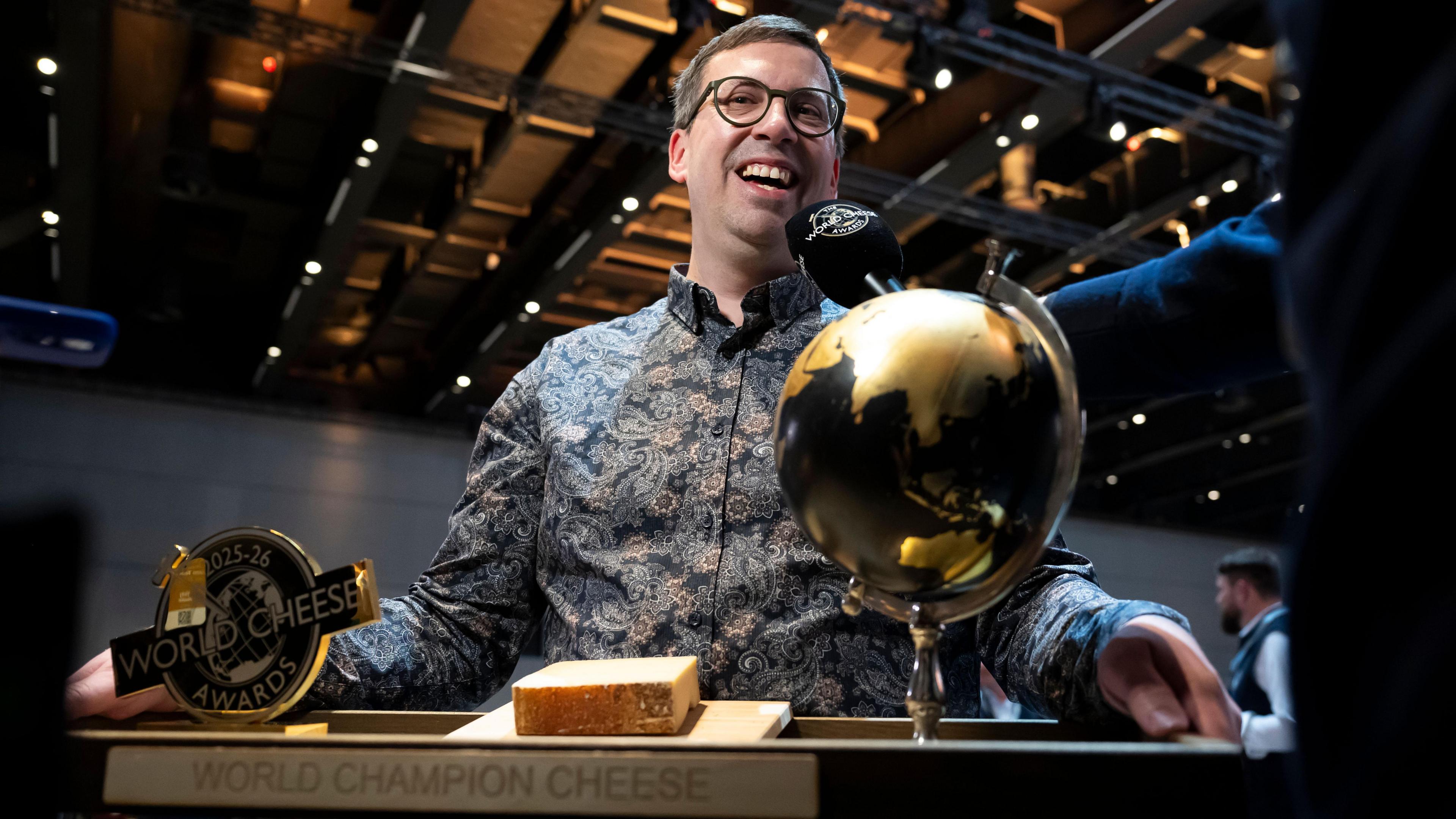 A man smiles as he holds a board with a globe trophy and a piece of cheese, with the title "World Champion Cheese" inscribed on