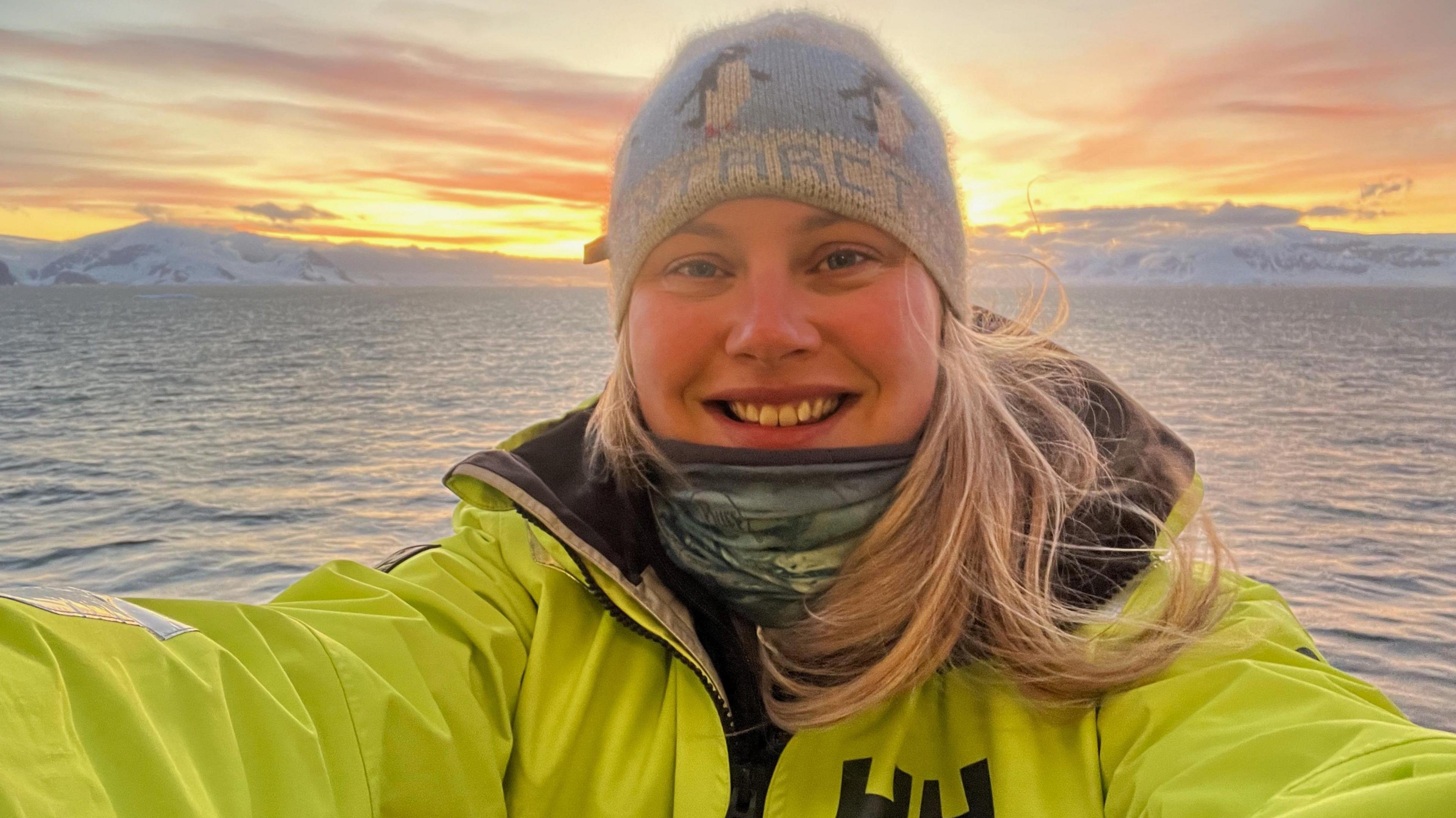 A woman on a boat in the ocean takes a selfie with a sunrise over glaciers visible in the background. She has long blonde hair and is wearing a blue and white hat with penguins on it, a green scarf and a lime green coat.