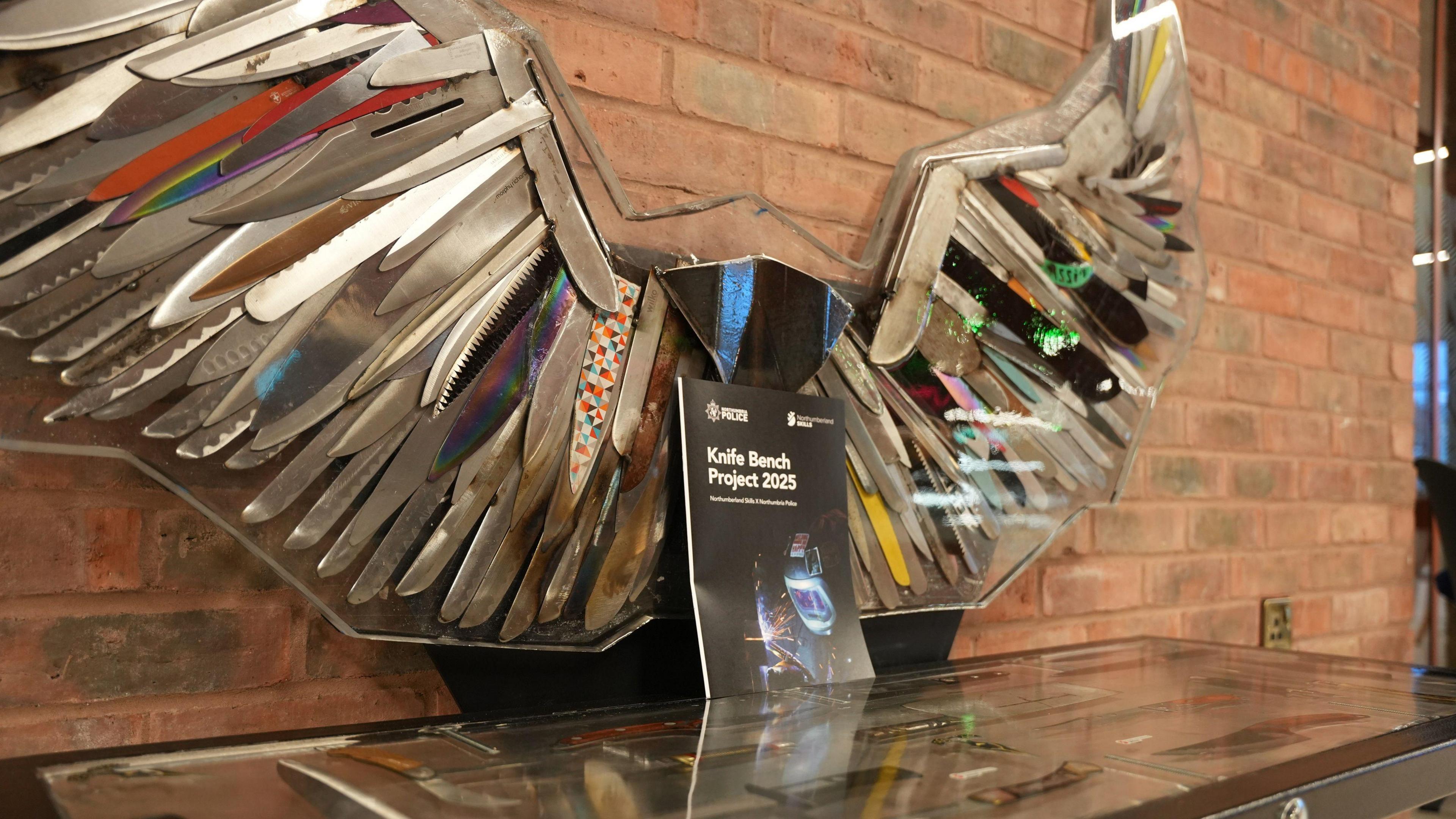 A close up on the bench, with a Knife Bench Project 2025 booklet rested on it. The back of the bench resembles the shape of spread wings, and has been created by various knives, of different colours. The majority are silver, but numerous other colours are also visible, including rainbow designs and also yellows and blacks.