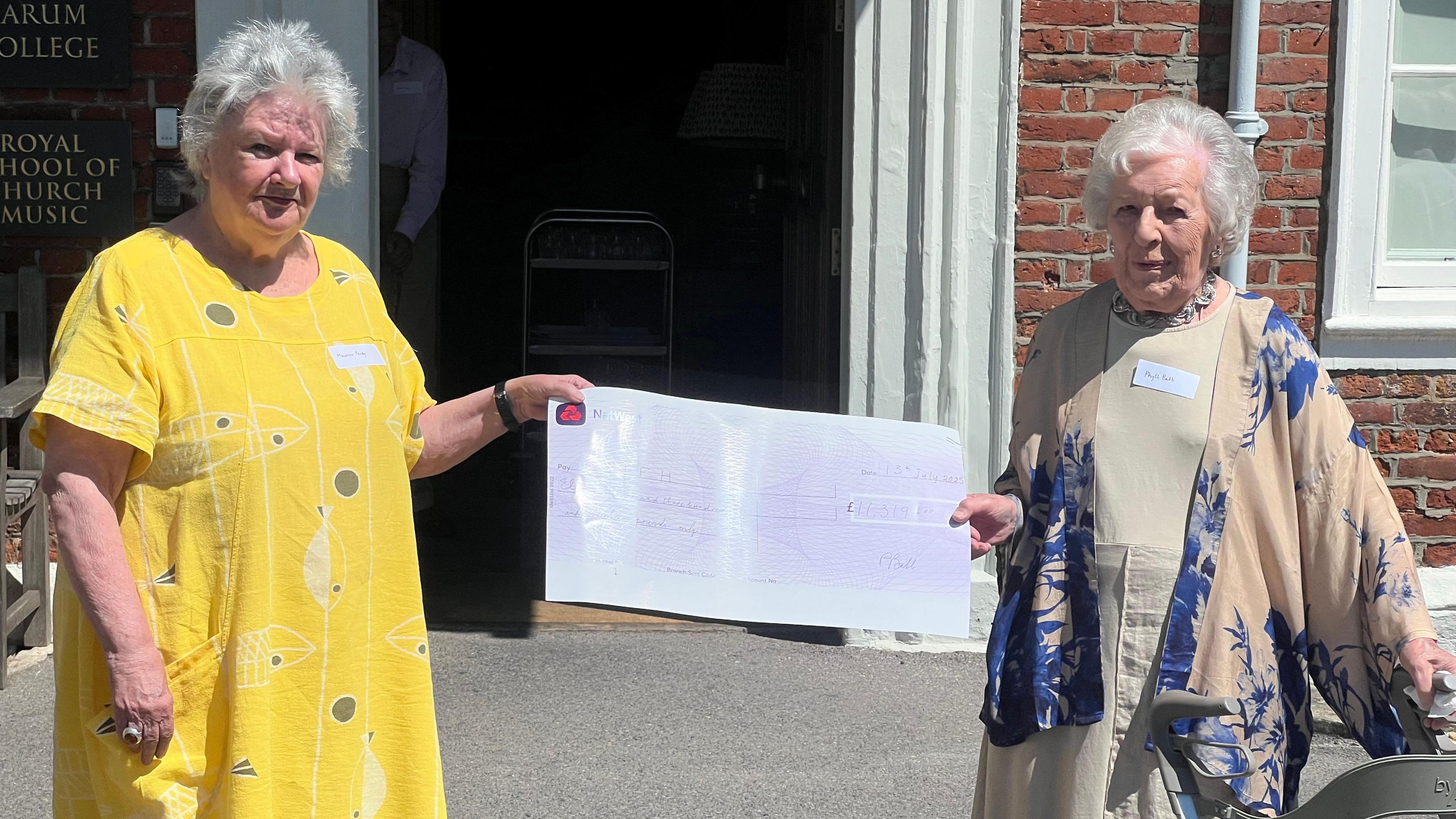 A woman in a yellow dress and short grey hair holds a large cheque that is also held by another older woman (Phyll Babb) who has short white hair and is wearing a beige and blue kimono style jacket and beige dress.