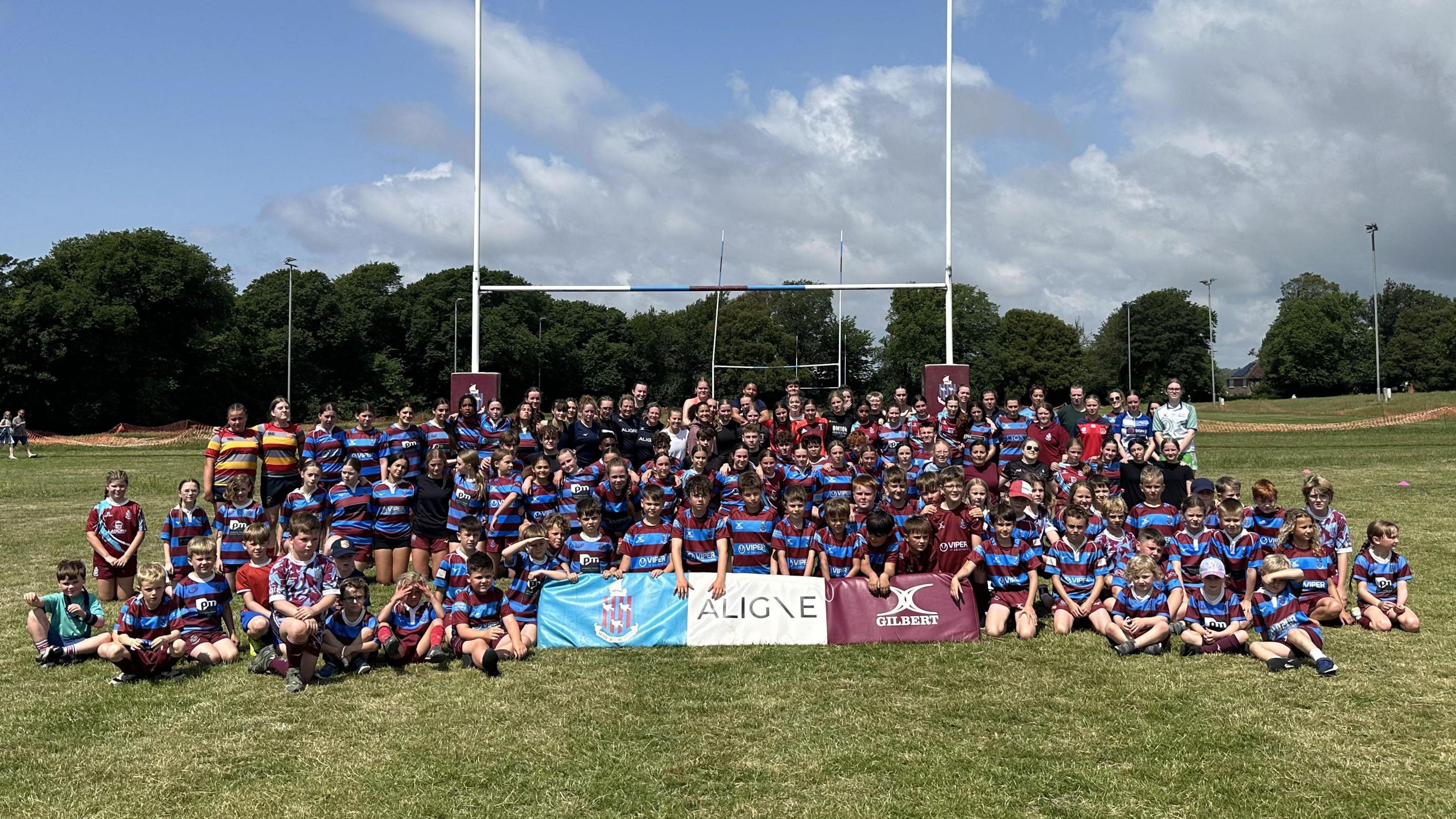 A group of young rugby players wearing blue and red shirts. They are stood underneath some rugby posts.