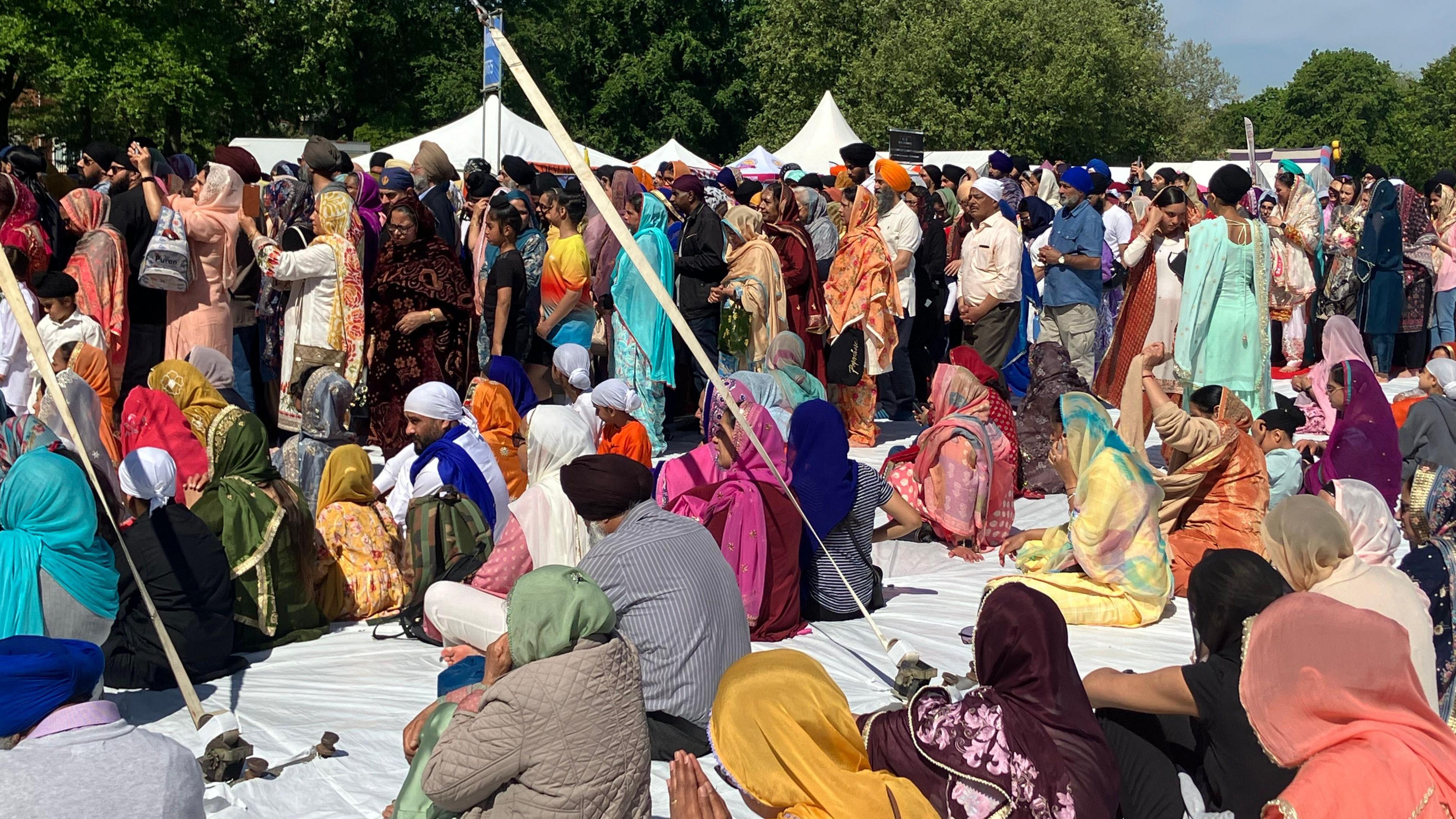  A group of more than 100 people dressed in traditional south Asian attire. The people closest to the camera are sitting on a white sheet on the ground while in the background a large group of people is standing.