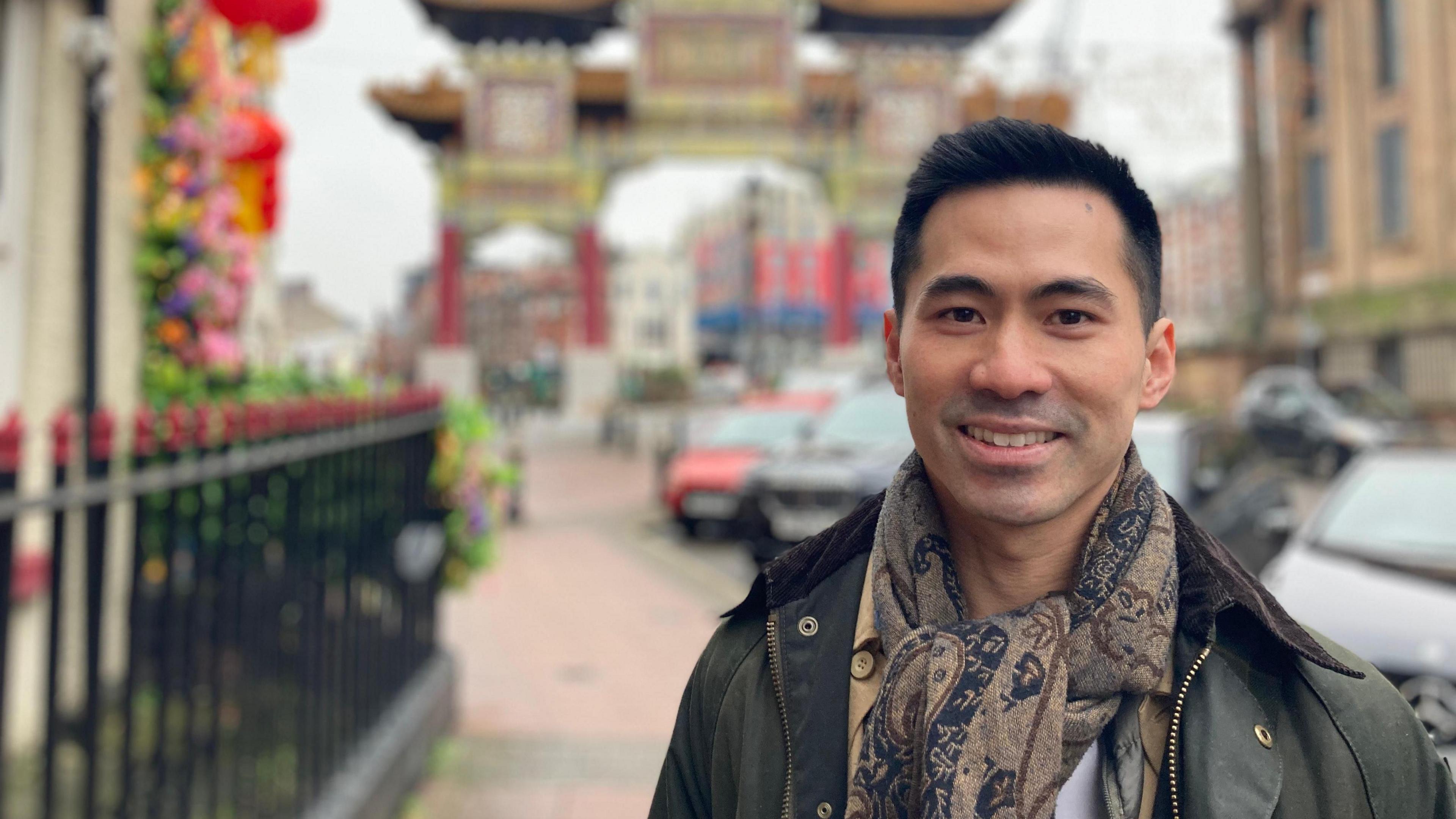 A young Chinese man smiles at the camera, wearing a jacket and scarf as he stands on Nelson Street 