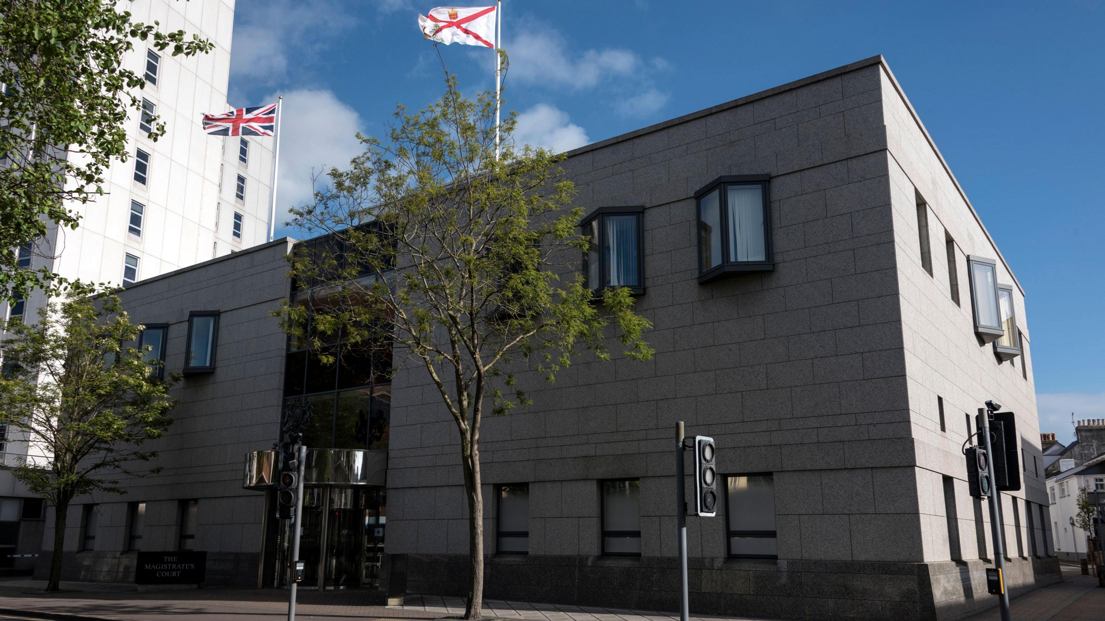 The exterior of the Magistrates' Court in St Helier which has grey walls and several windows. The Union flag and Jersey's flag are flying at the top of flag poles. It is a sunny day.