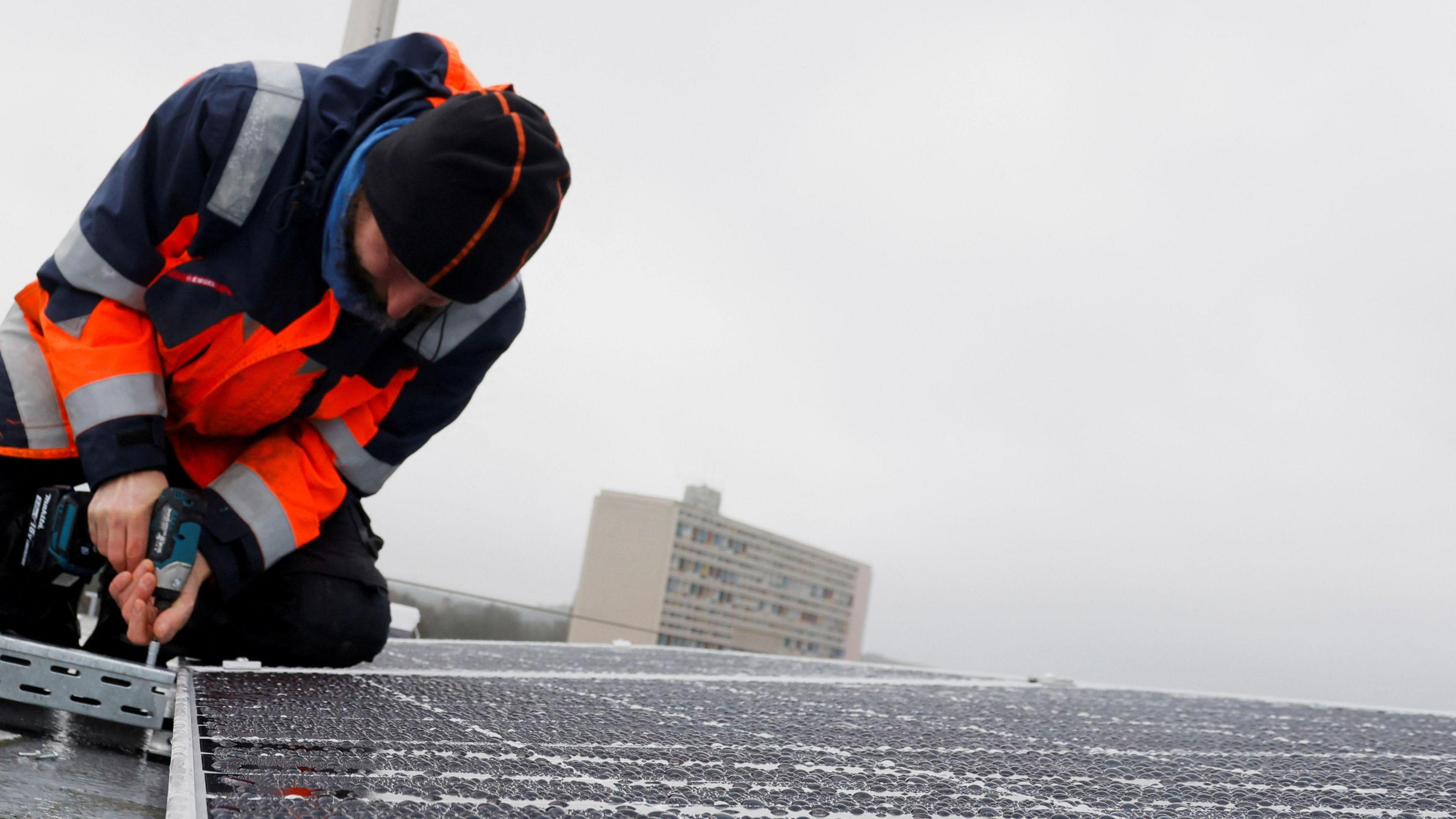 A stock image of solar panels being installed on a roof. An engineer wears a hi vis jacket and uses a drill. 