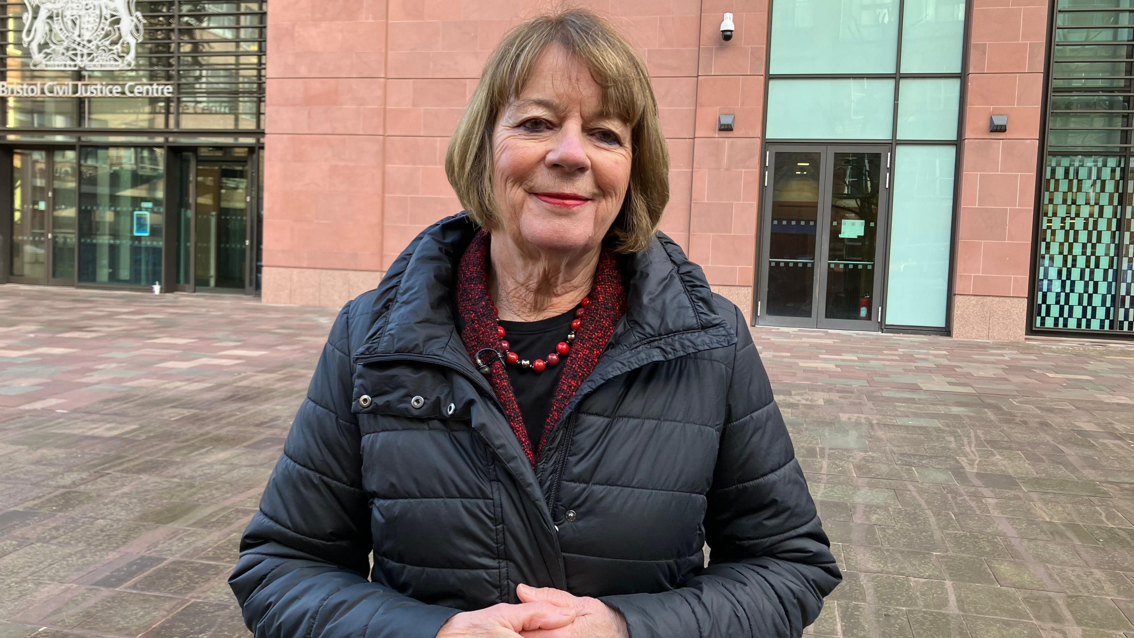 Sandra Fryer, chair of governors at Cotham School standing with her hands clasped together in front of a court building. She is wearing a dark coat, red scarf and is smiling at the camera