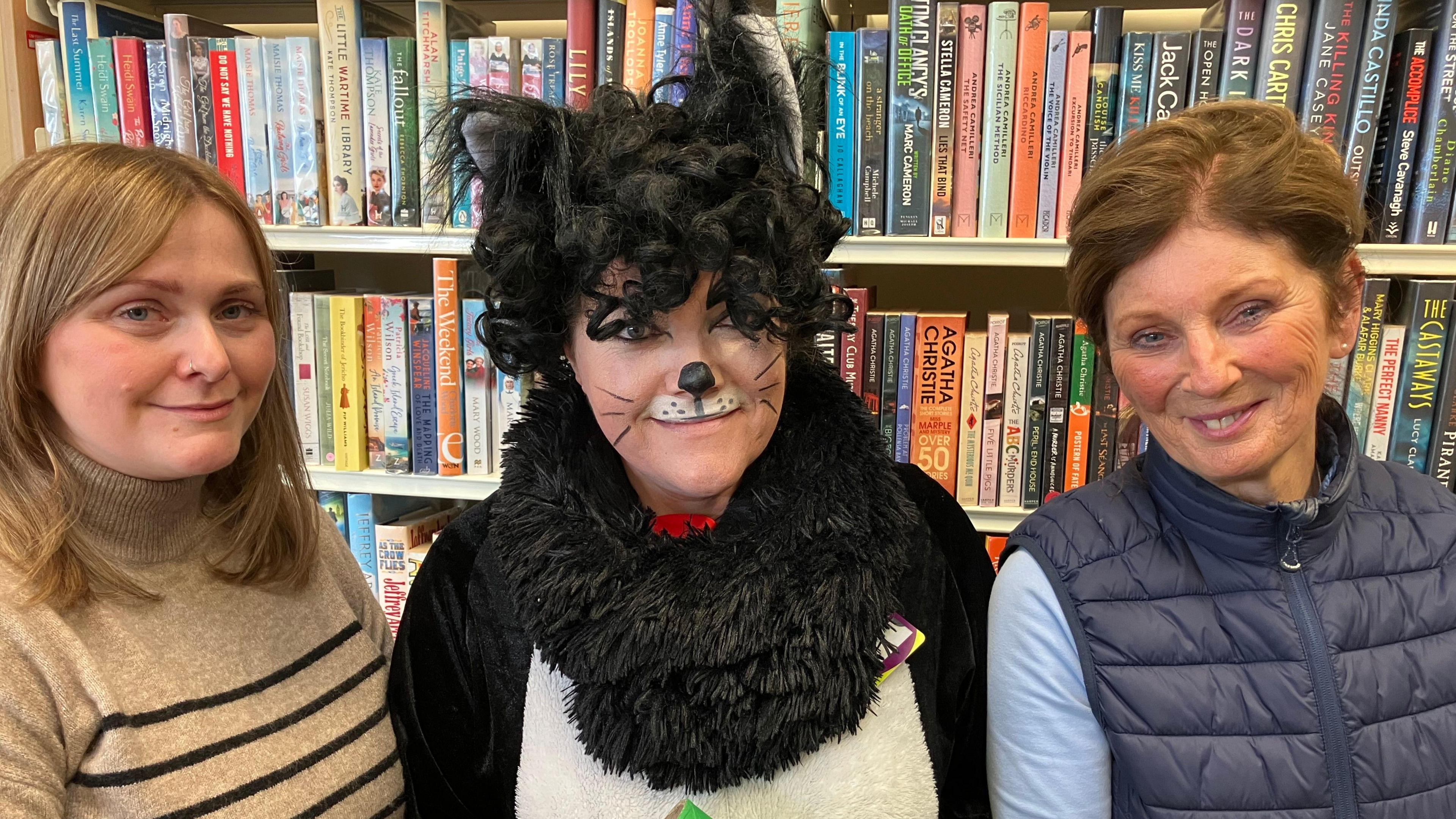 Vicky Fedak who has long light brown hair, wearing a beige and black striped top with the Library Cat who is dressed up as a cat with black and white fur and her face is painted with black whiskers and nose. Carol is standing to the right and has brown hair that's tied back, she's wearing a navy gilet and blue top. They are all smiling and standing in front of a bookcase full of books. 