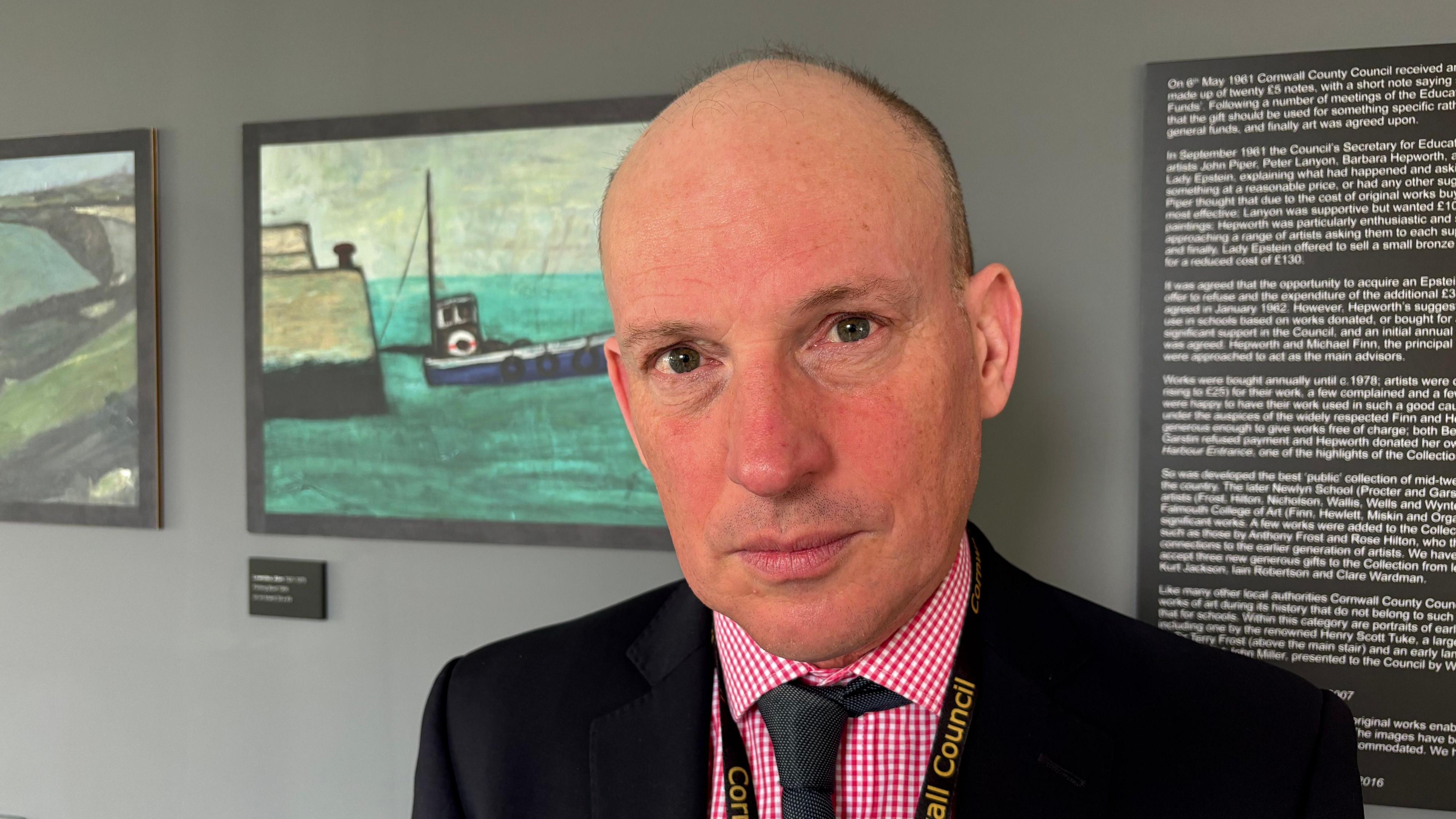 Counsellor Loic Rich. He is looking into the camera. He has a bald head and is wearing a black suit and tie with a red and white gingham shirt. 
