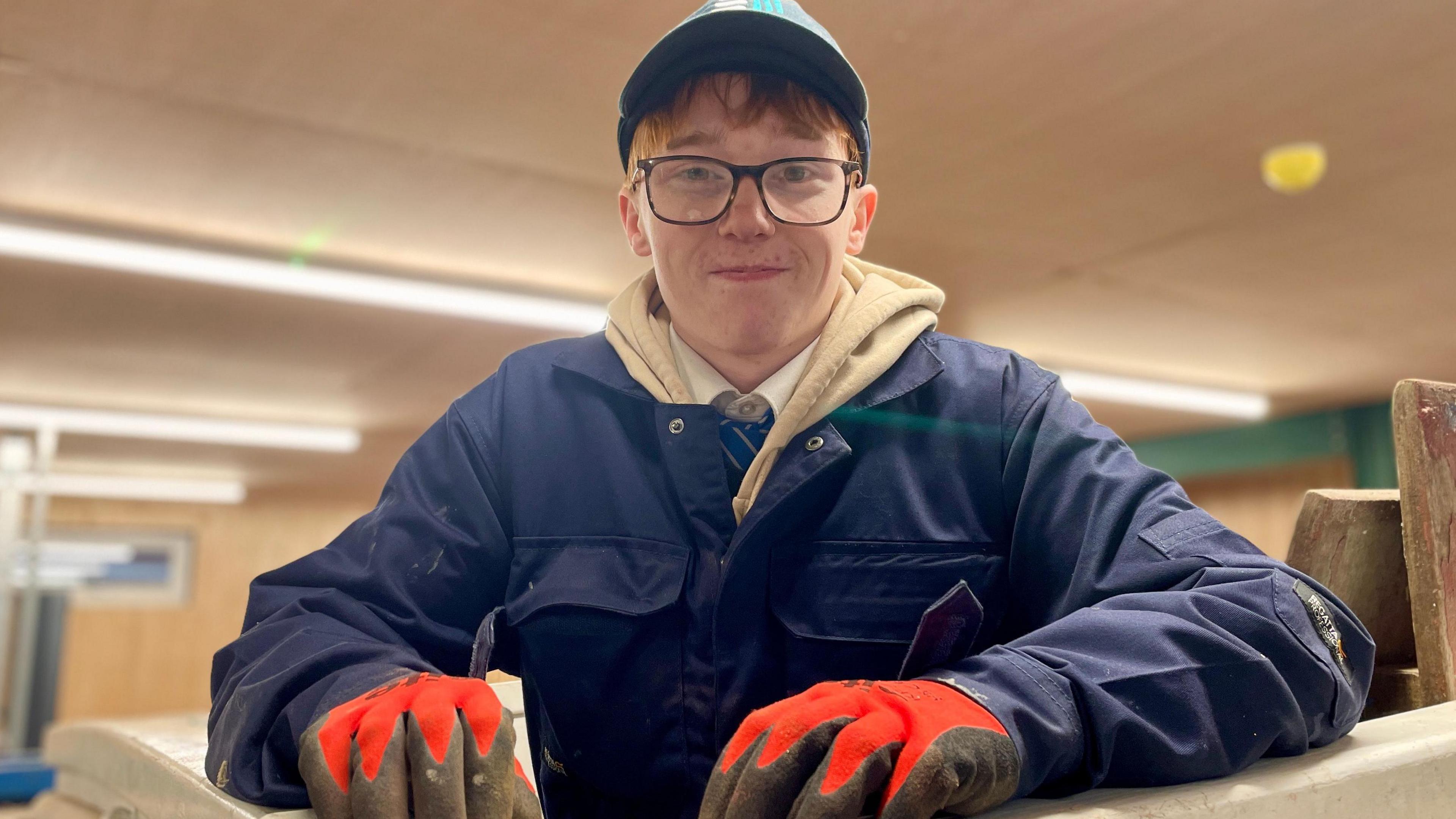 A teenager wearing glasses,  a cap, blue overalls and safety gloves leans over a bench,  facing the camera. 