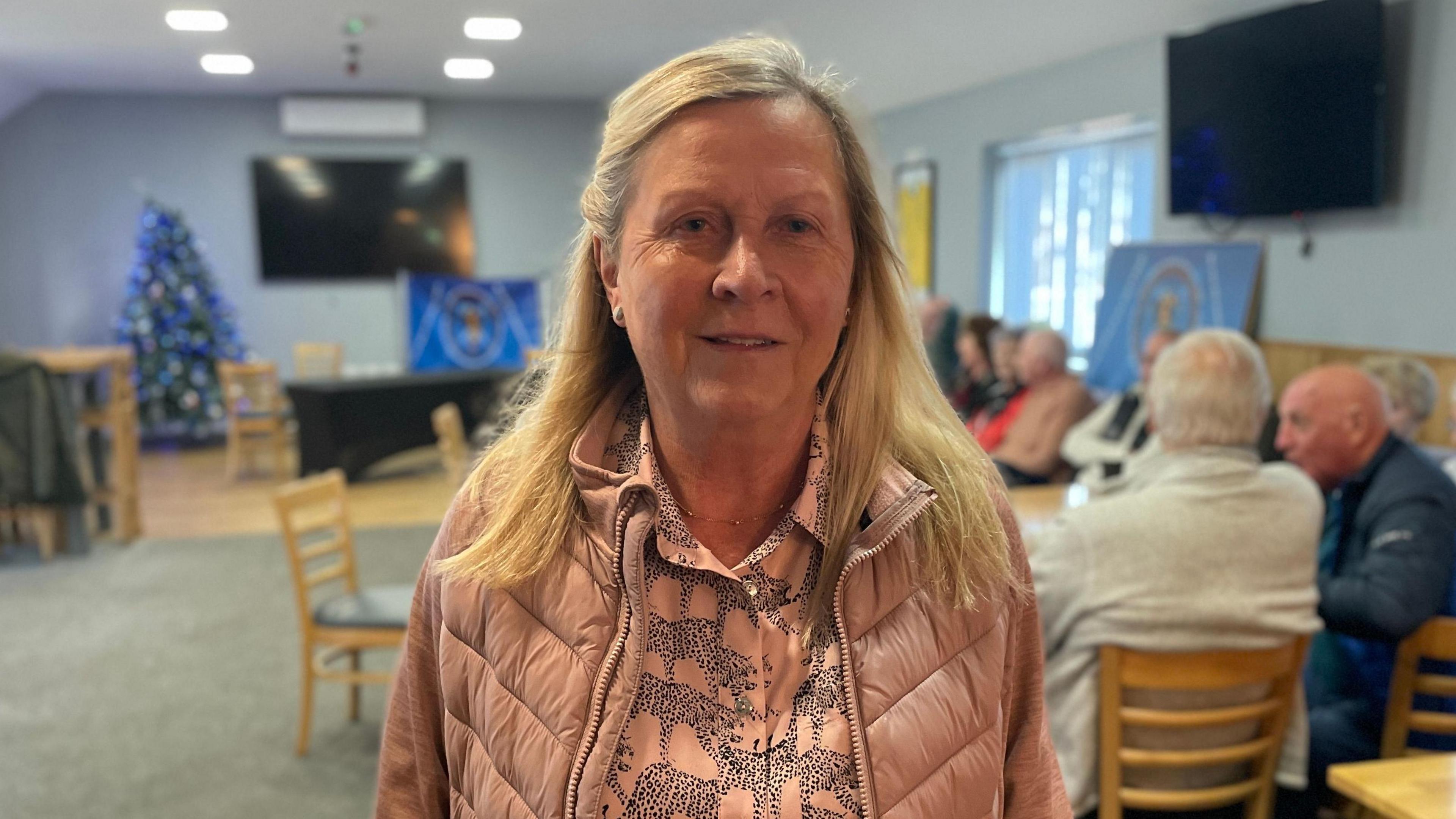 Kim Smith, a middle-aged woman with long blonde hair and wearing a pink padded winter jacket and pink and back patterned blouse. She is standing in a room with several sets of tables and chairs and several people seated in the background.