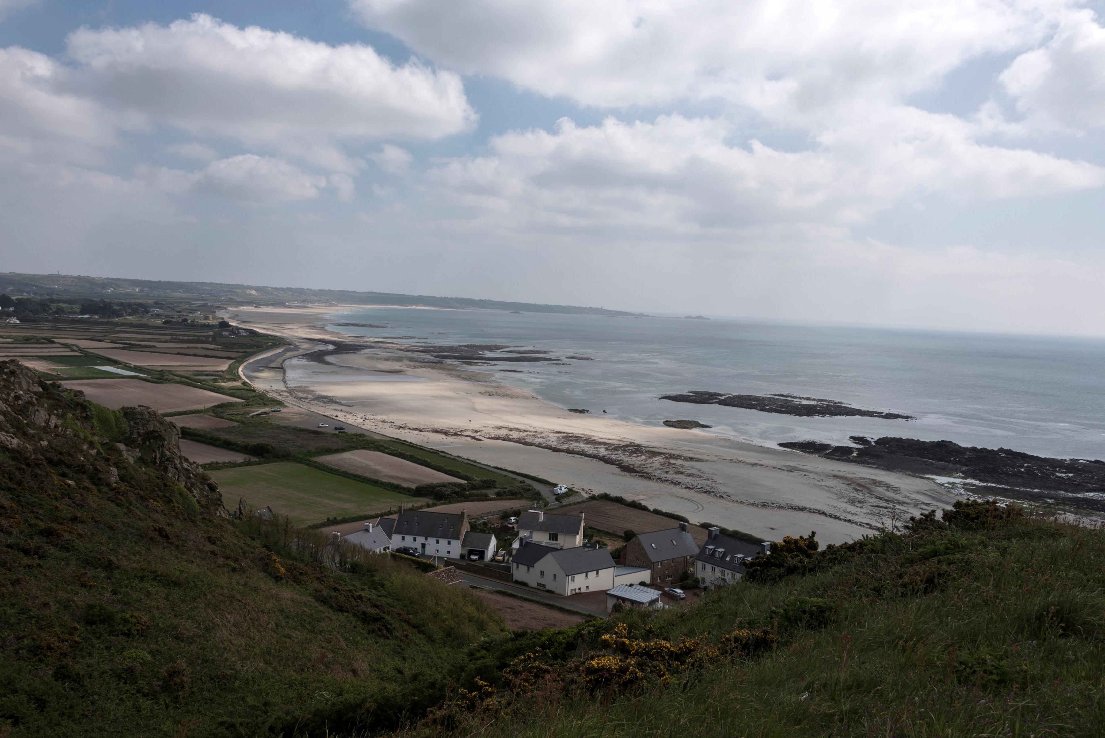 A wide double bay with an expanse of cream sand and some rocks exposed.  The cliffs are green with some yellow gorse, There is a small cluster of white buildings with dark roofs in the foreground.