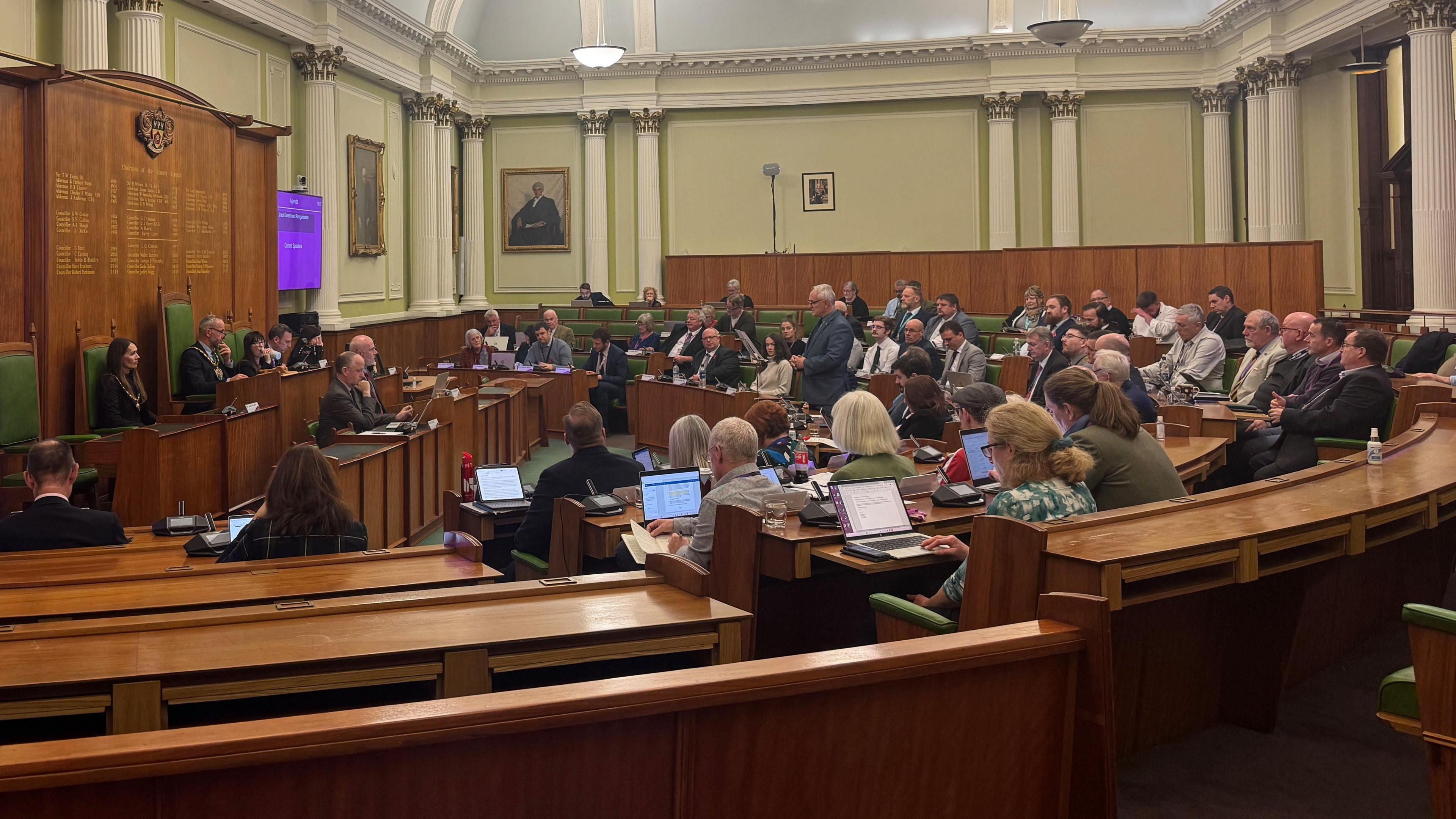 The Derbyshire County Council chamber with Councillors in a horseshoe range of seats to the right of the picture. The Chairman and Officers on a raised dais to the left of the image.
Leader Alan Graves is standing to speak.