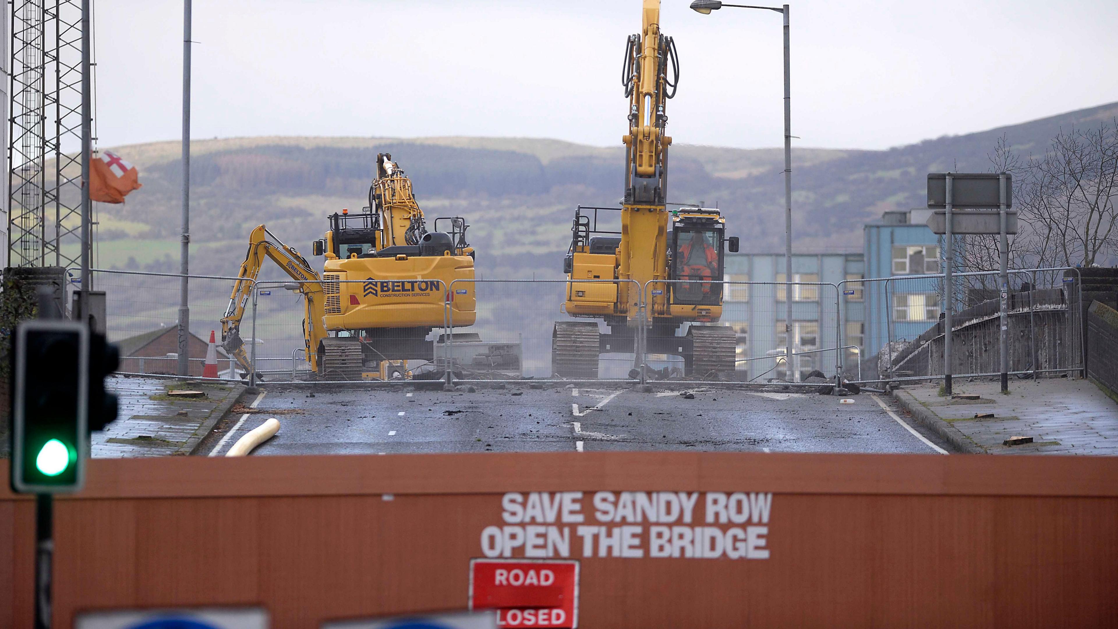 Two large diggers ripping up a road. The road is bloacked by a barrier on which someone has written "Save Sandy Row, open the bridge". There is a Road closed sign in front of that. In the background some hills. 