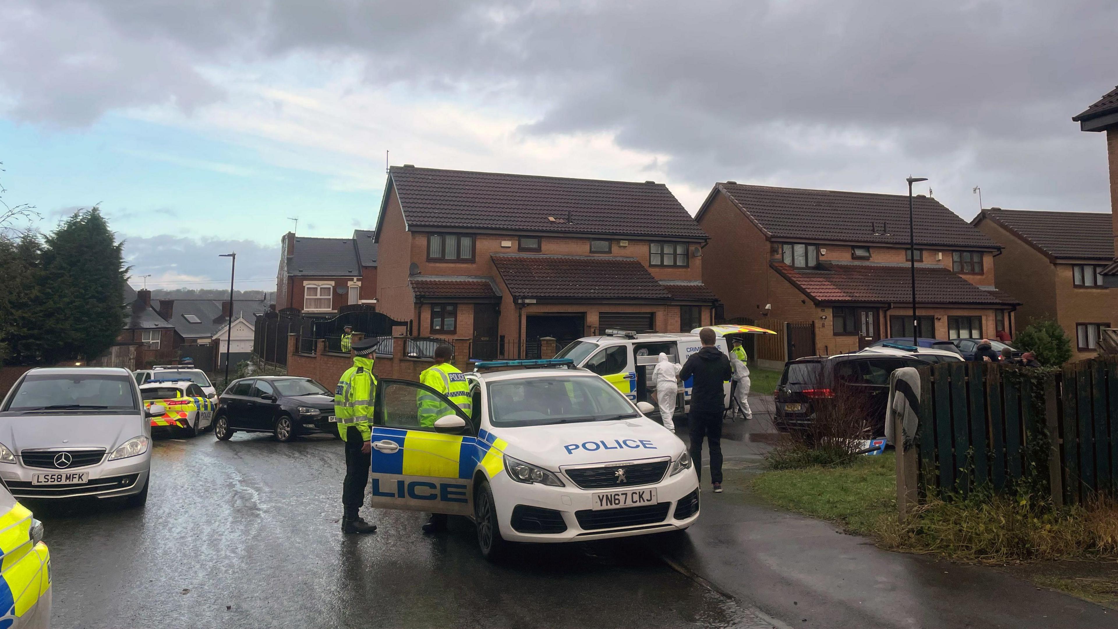 Several police cars and officers can be seen in the road on what appears to be a damp day. A crime scene investigations van is parked outside a property.