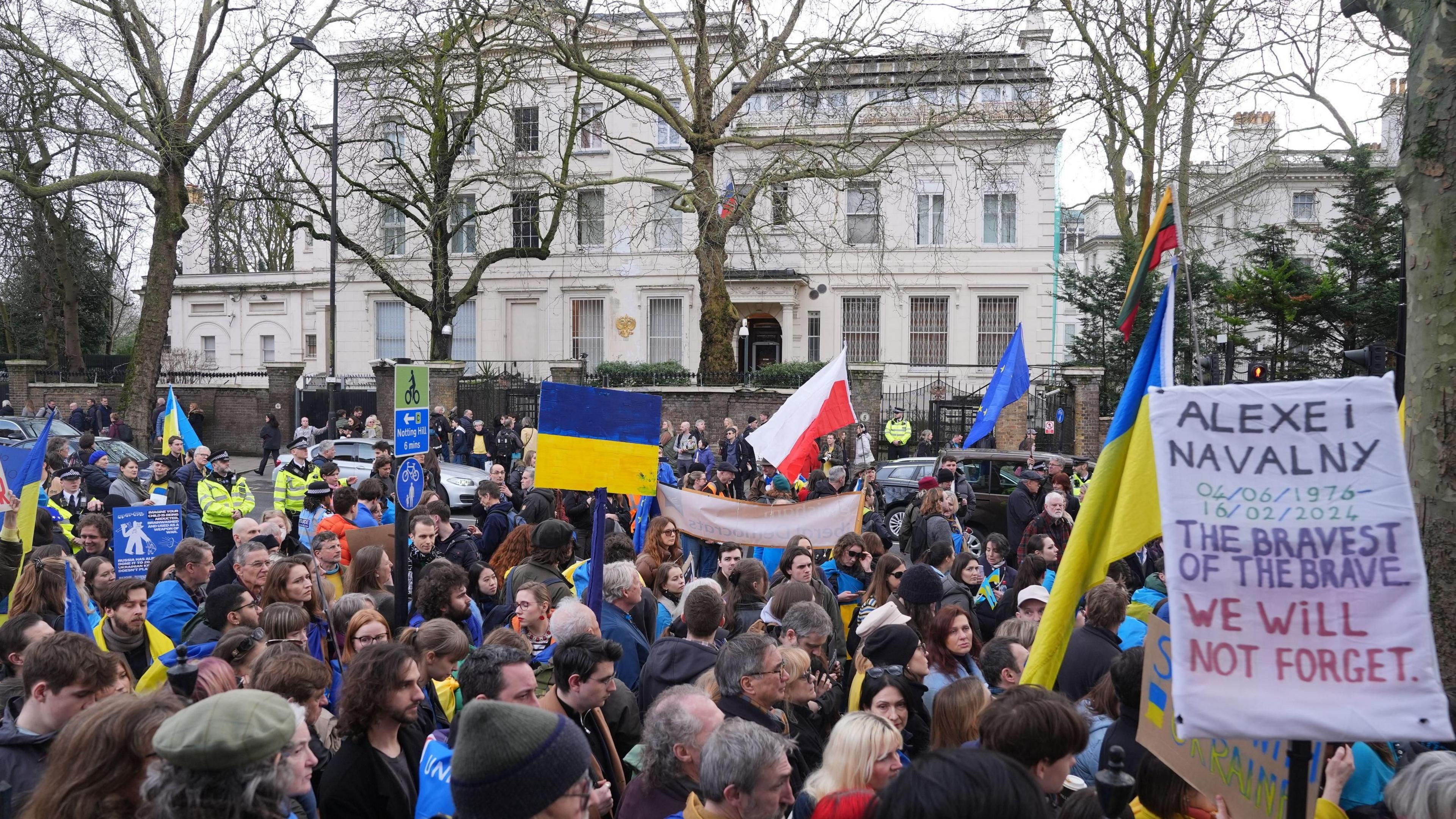 A group of protesters - holding Ukraine flags and a poster reading "Alexei Navalny 04/06/1976-16/02/2024 The bravest of the brave, We will not forget - outside the Russian embassy in London - a grand white building with a Russian flag flying over it's door.