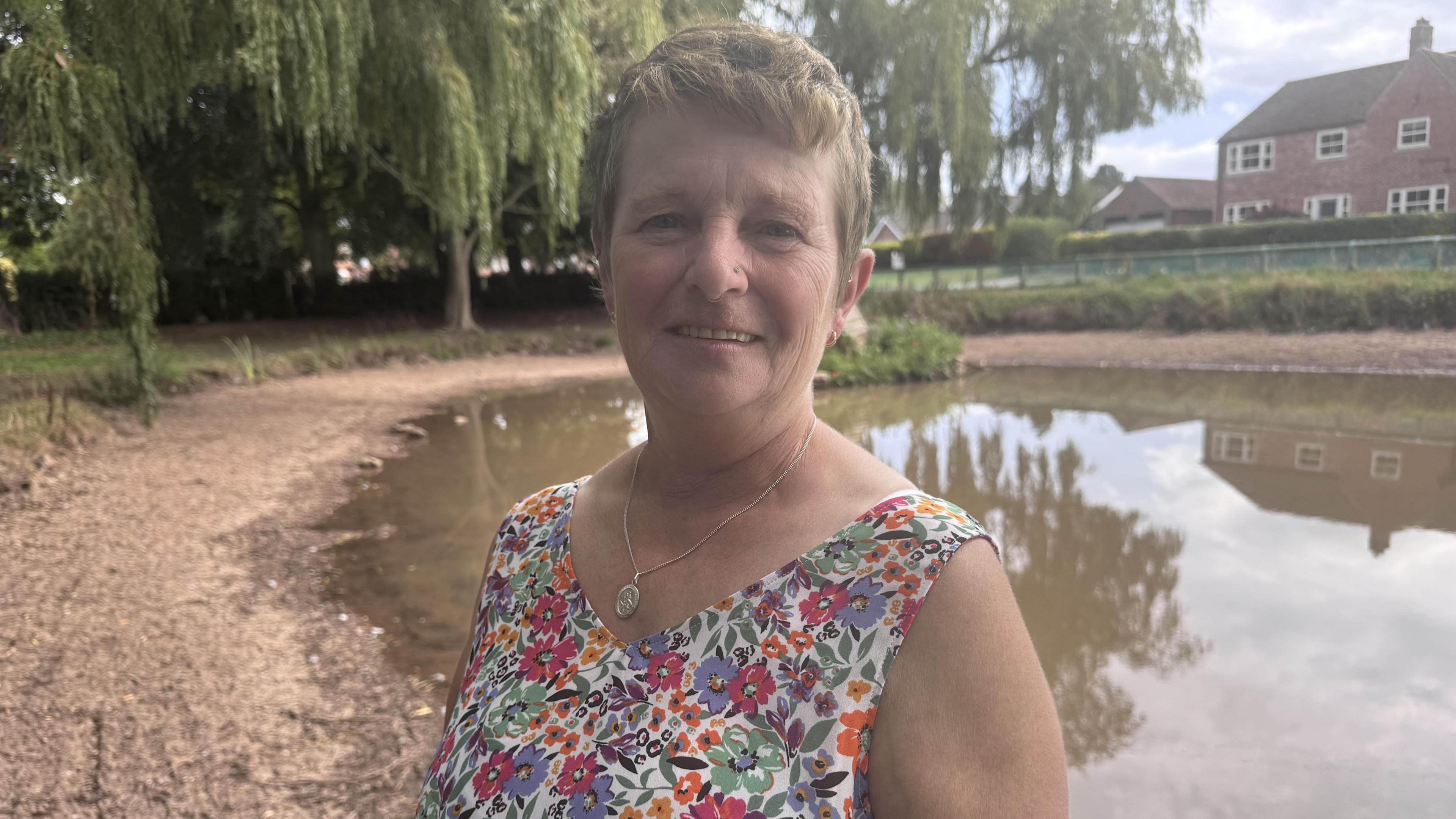 A woman with short hair and a colourful flowery vest top, with a silver necklace. In the background is a pond.