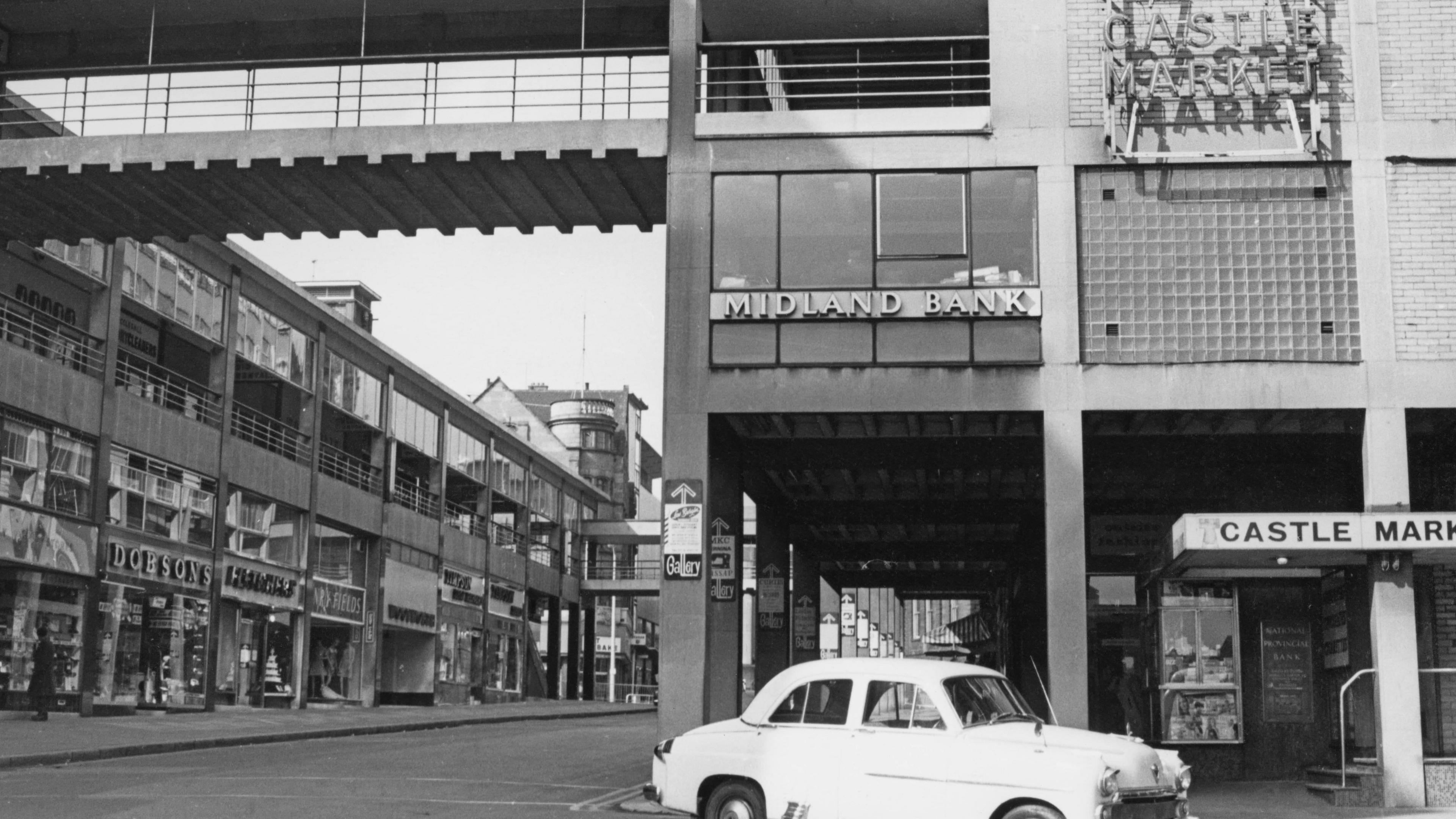 A 1960s photo of a city street, with a small white car in the foreground and an empty road in the left-hand background 