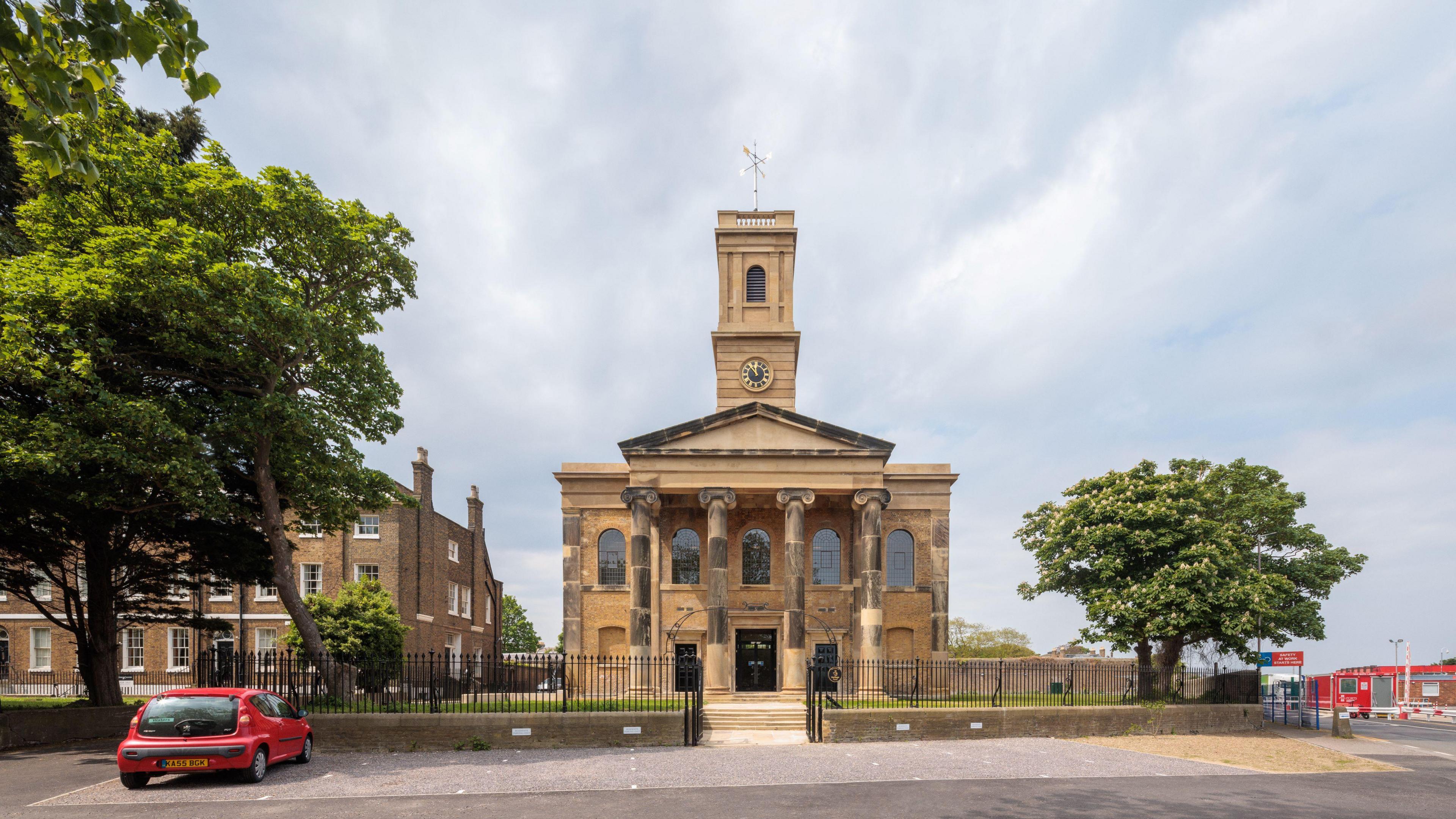 A church building, centre, surrounded by trees. A red car is parked in a car park in front of the church. The photo is set against a blue, cloudy sky.