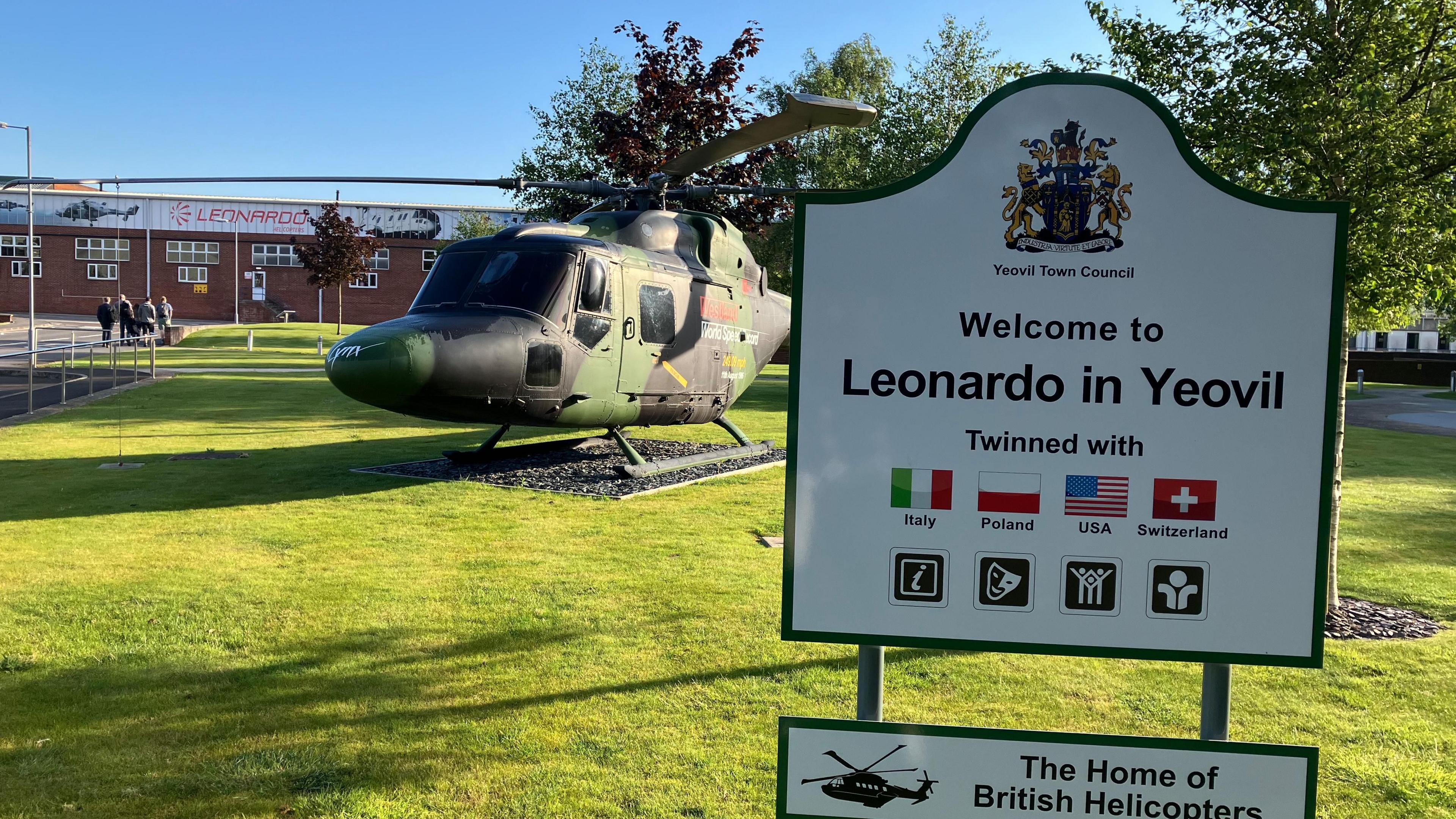 a sign reading 'welcome to leonardo in yeovil' in front of a helicopter sitting on a grass lawn