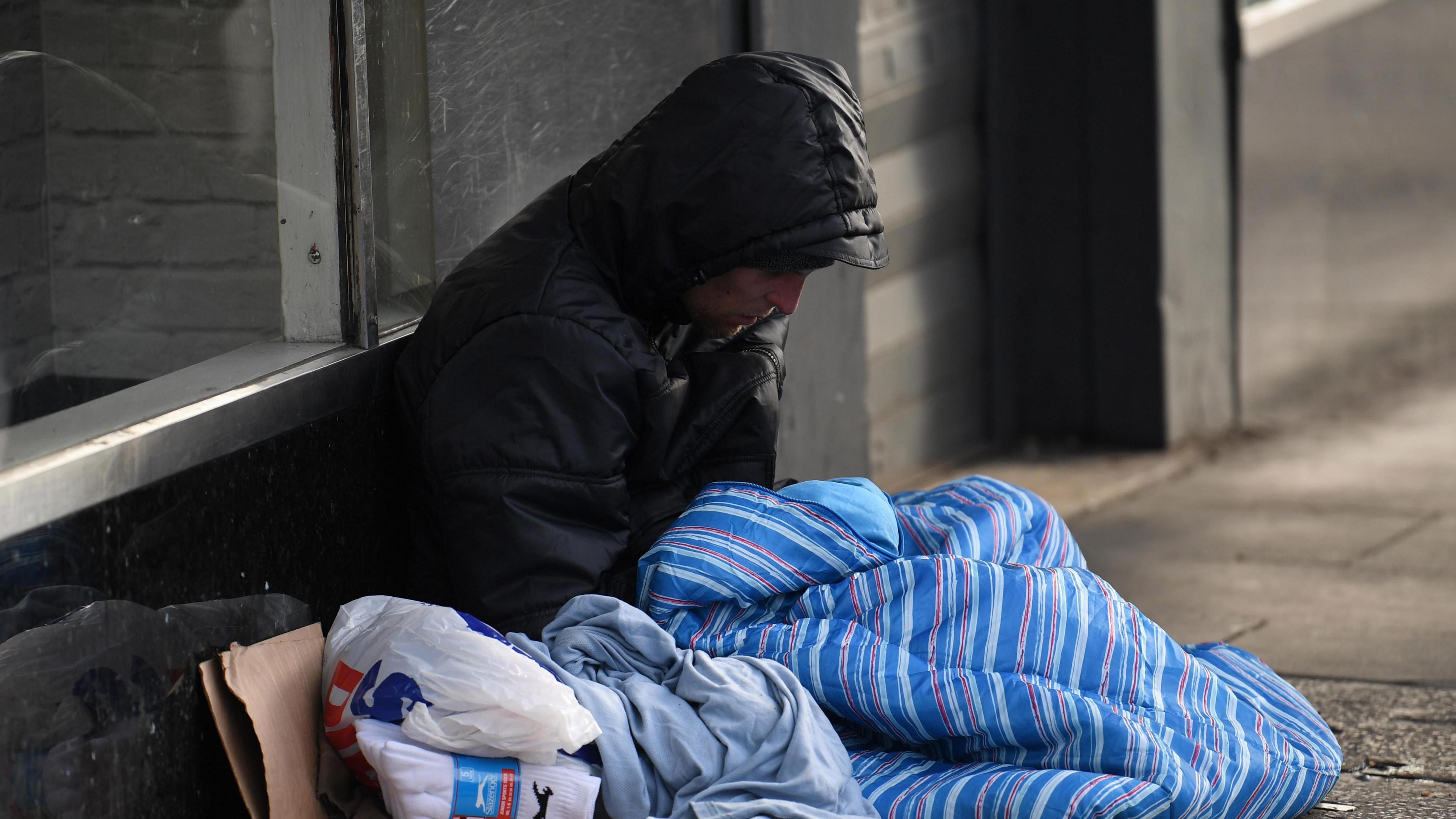 A homeless man with his back to a wall, sitting on a street. He's wearing a dark grey hooded jacket. He has a blue striped duvet blanket and a bag with some unopened socks.