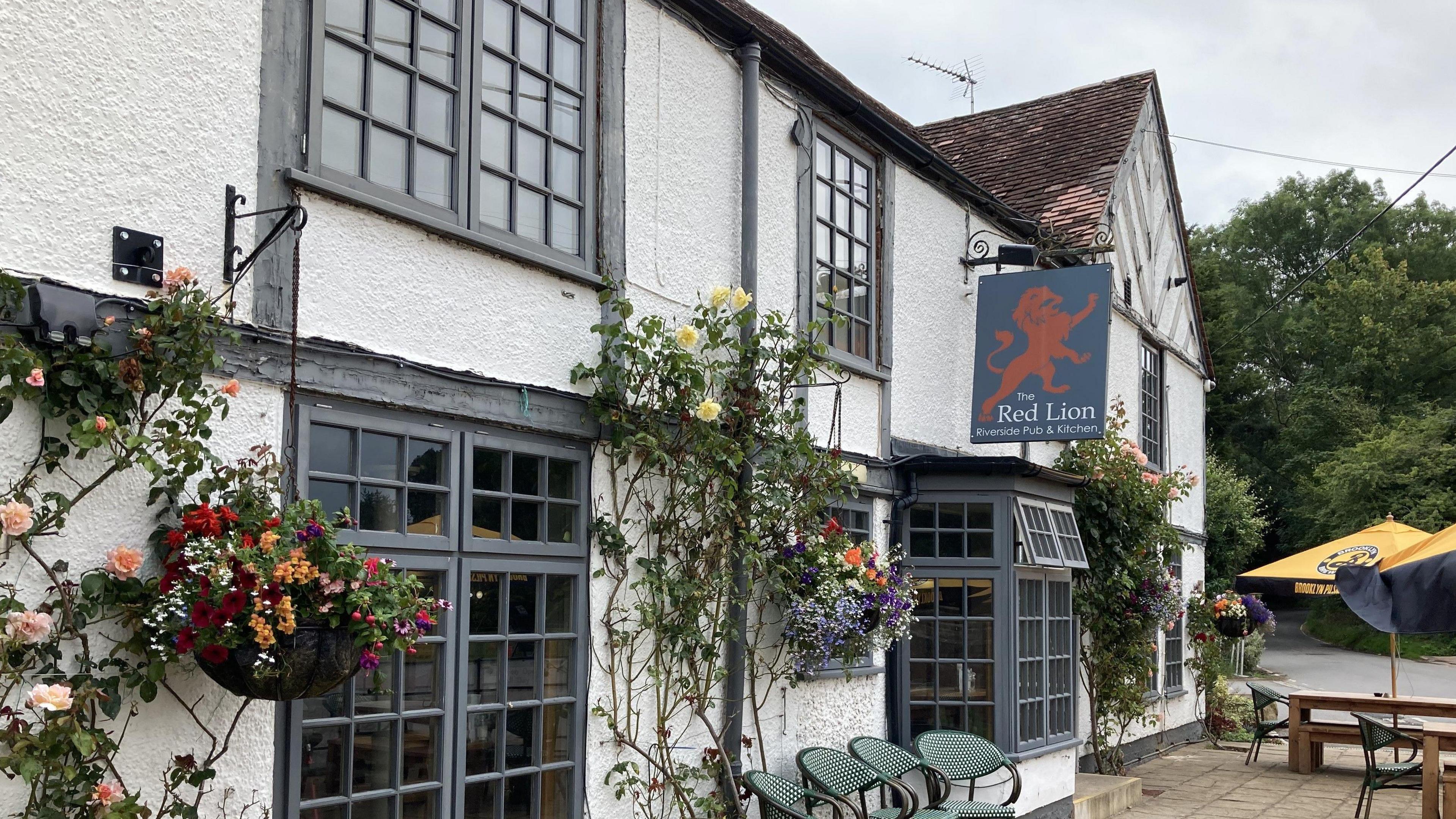 The outside of a pub with roses growing against a white wall as well as hanging baskets and benches and parasols on a terrace.