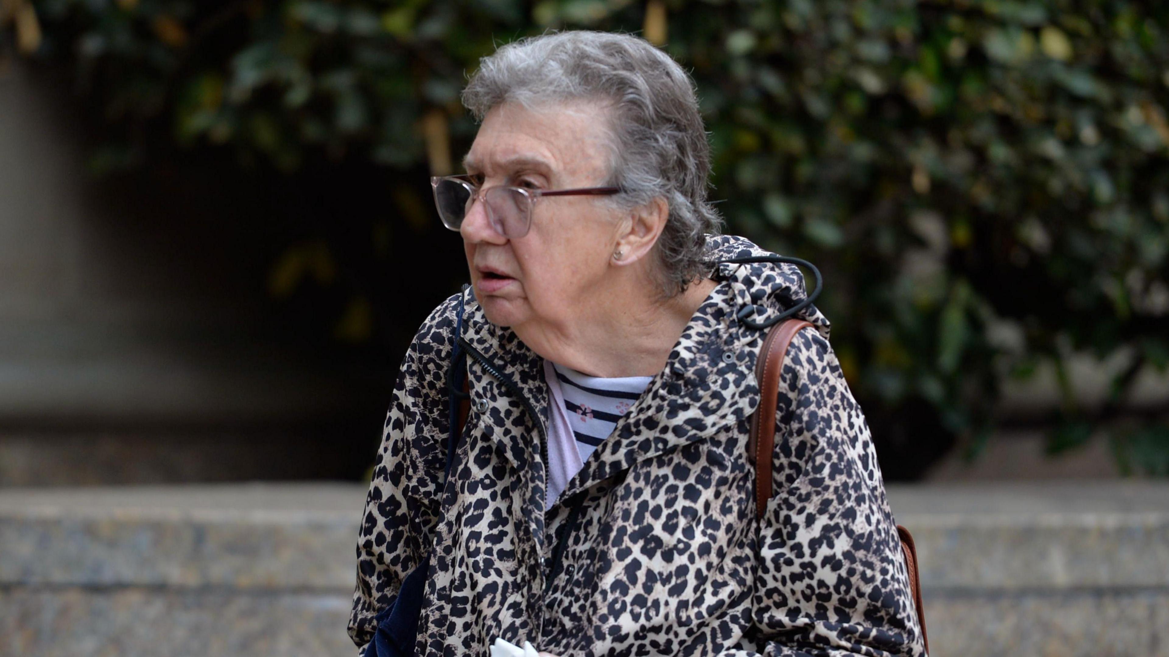 Patricia Robertson, a 77-year-old woman with short grey hair and clear-rimmed glasses, is walking right to left. She is wearing a leopard skin hooded jacket and a light and dark blue striped shirt underneath. She has a brown leather bag slung over her shoulder.